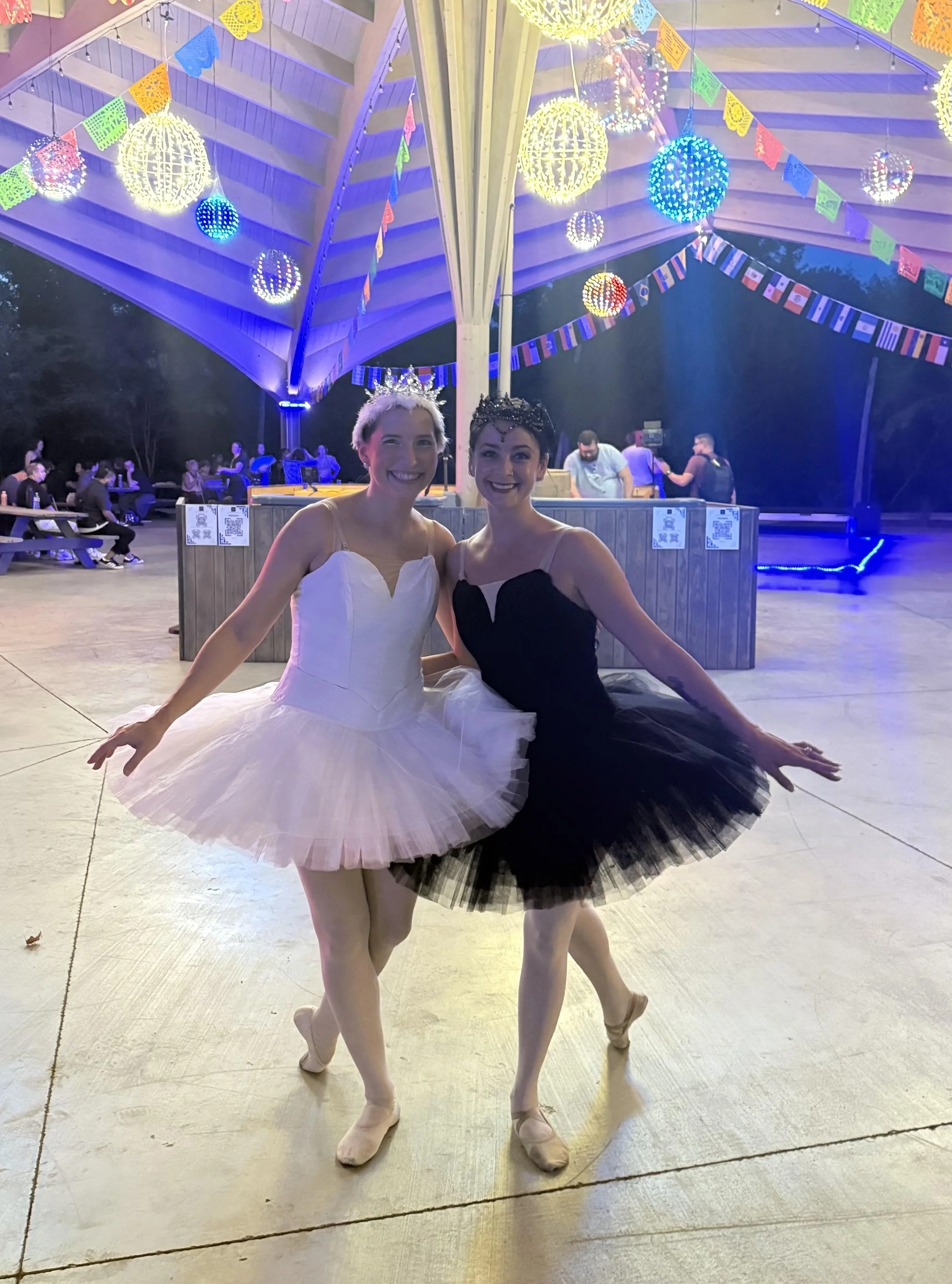 Two ballet dancers in tutu costumes, one in white and one in black, smiling and posing together in front of a decorated pavilion with colorful papel picado and hanging spheres during a festival or celebration.