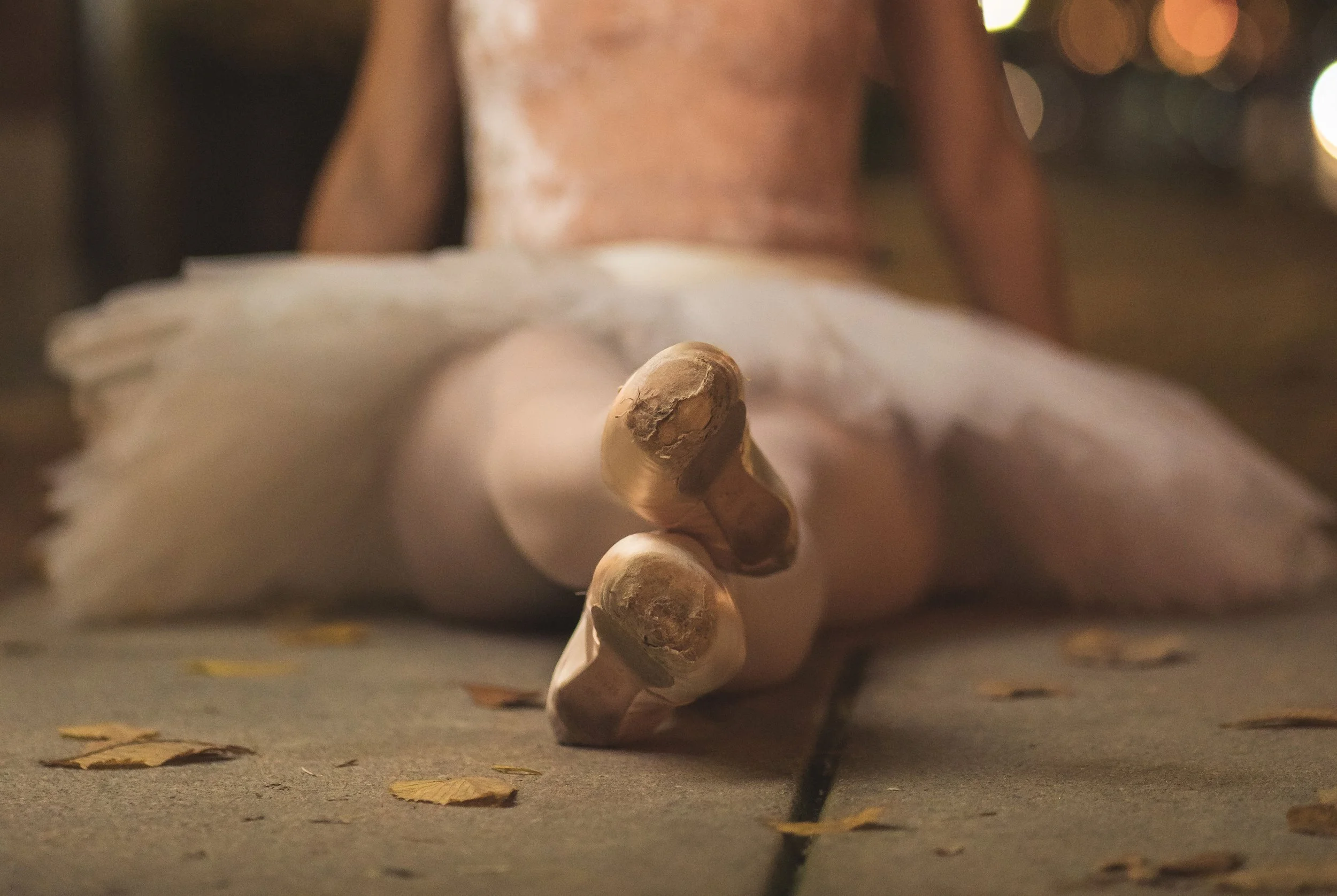 Close-up view of a person's pointe shoes over fallen autumn leaves on the ground at night, with a blurred background of city lights.