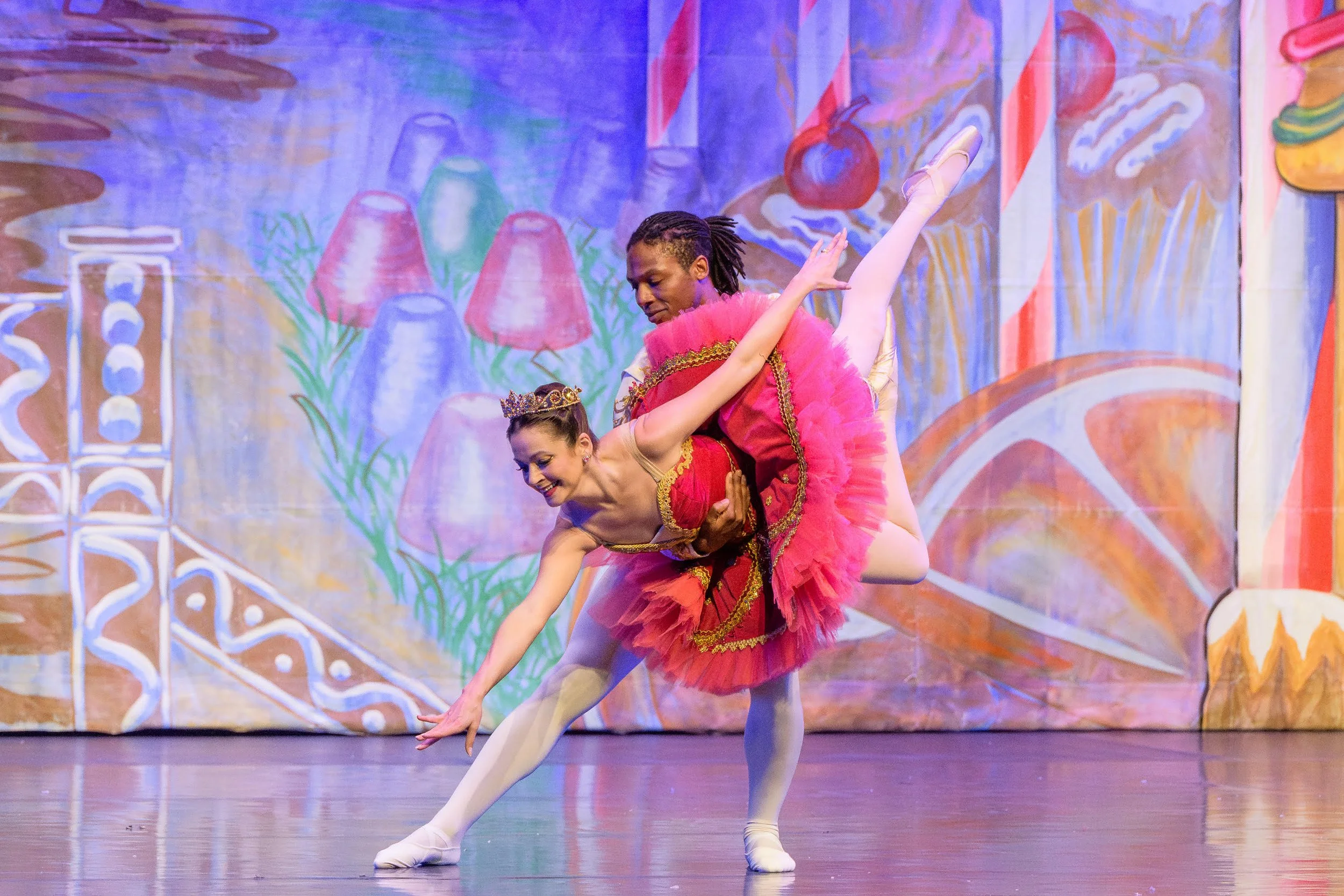 Two ballet dancers perform on stage, with one dancer dressed in a red costume and the other in a pink tutu, against a colorful painted backdrop.