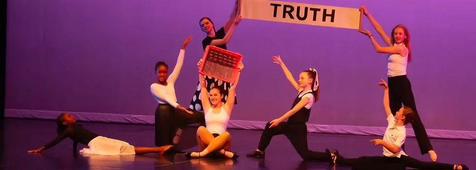 Group of children and a woman holding a banner with the word "TRUTH" during a dance or performance on stage with purple background.