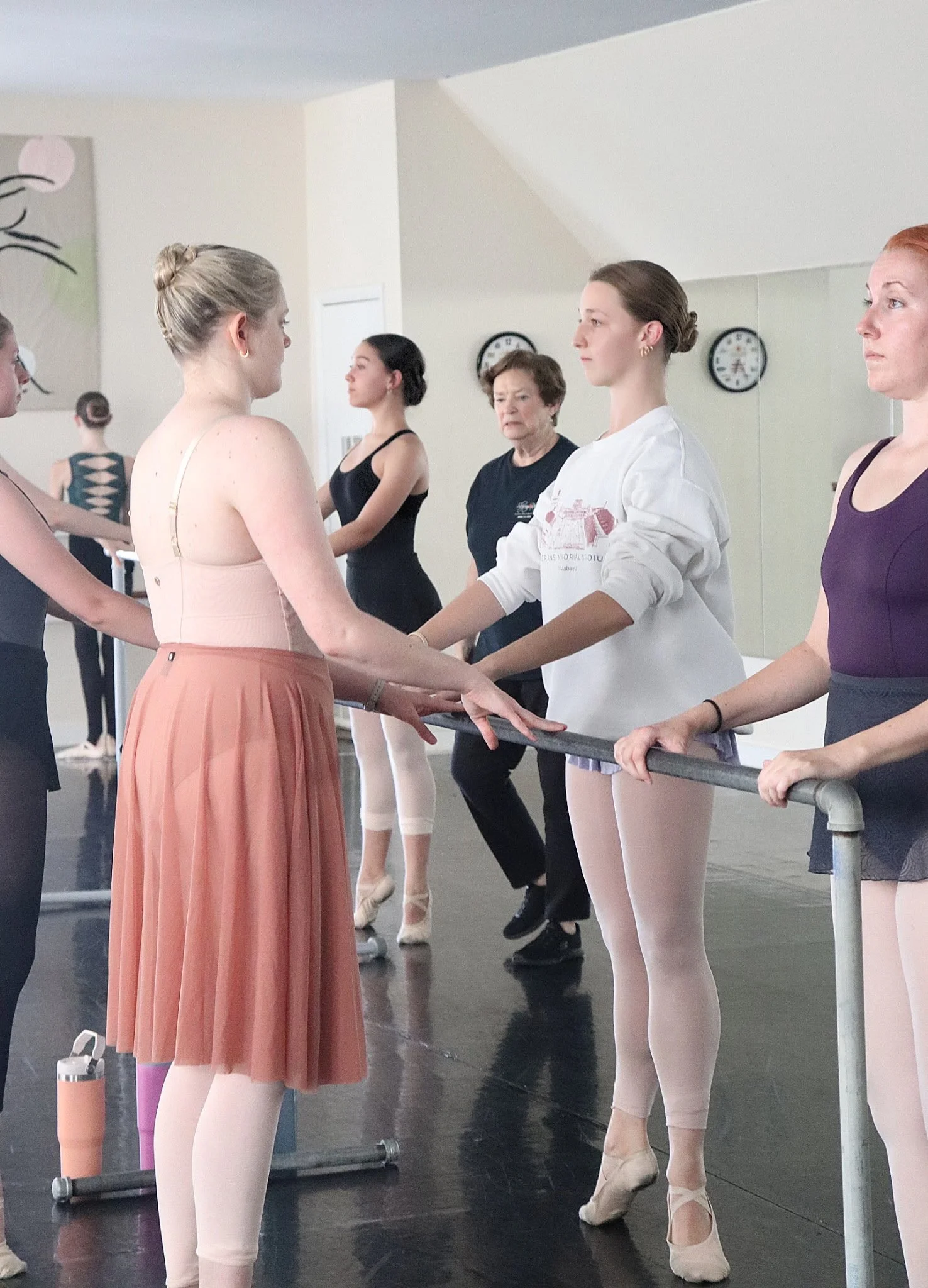 Ballet dancers practicing at a dance studio, holding hands at the barre, wearing ballet attire and pointe shoes, with mirrors and clocks on the wall.