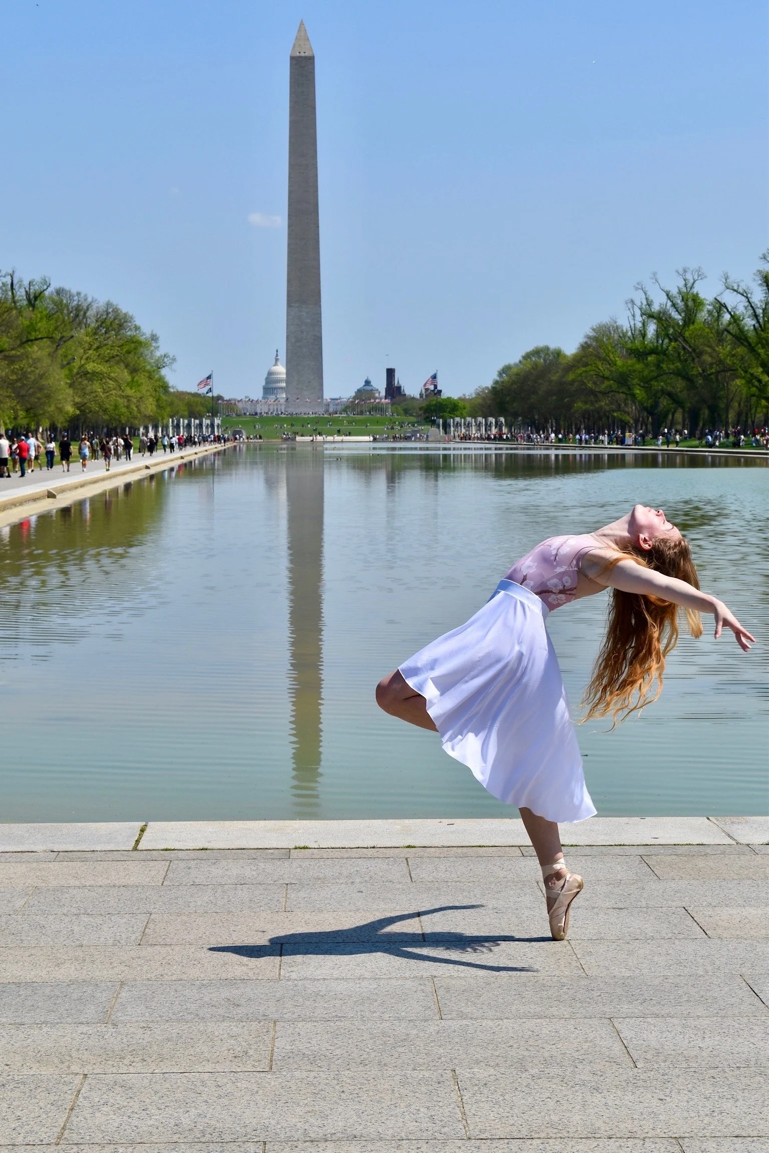 A woman in a white dress and ballet shoes dancing ballet on a stone pathway near the Reflecting Pool with the Washington Monument and U.S. Capitol in the background on a clear, sunny day.