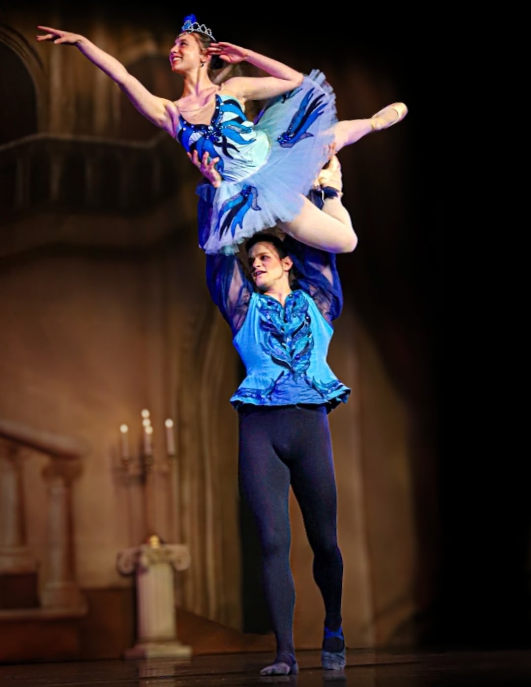 Ballet dancers performing on stage, with one dancer balancing on the other's shoulders, wearing blue costumes, in a theater with wooden architecture.
