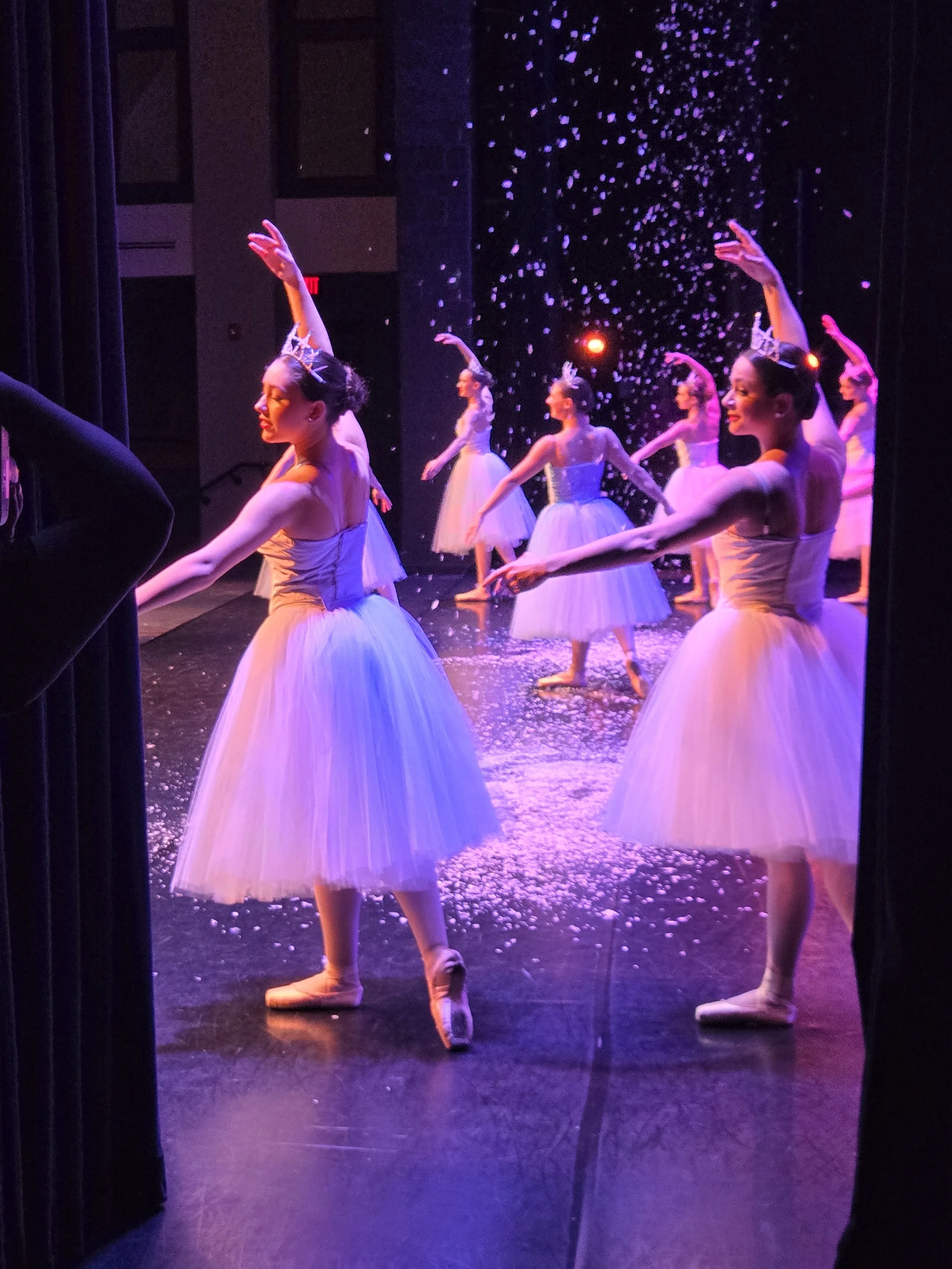 Ballet dancers are performing on stage with confetti falling, illuminated by purple and orange lighting.