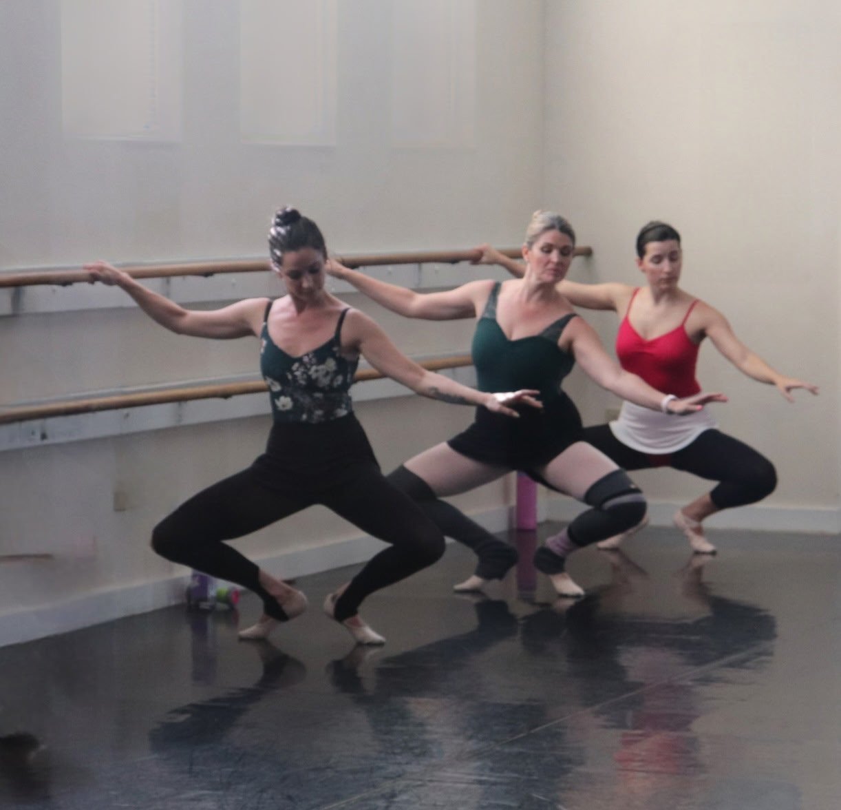 Three women practicing ballet at a barre in a dance studio.