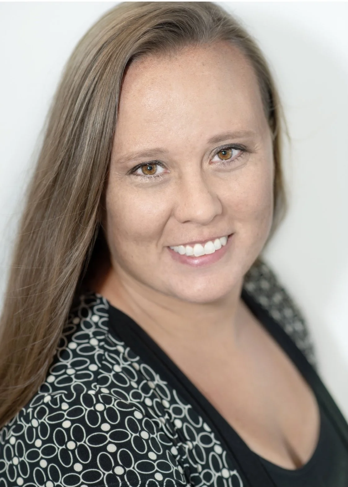 Close-up portrait of a smiling woman with long brown hair, wearing a black top with a patterned outer layer with circular designs, against a plain light background.