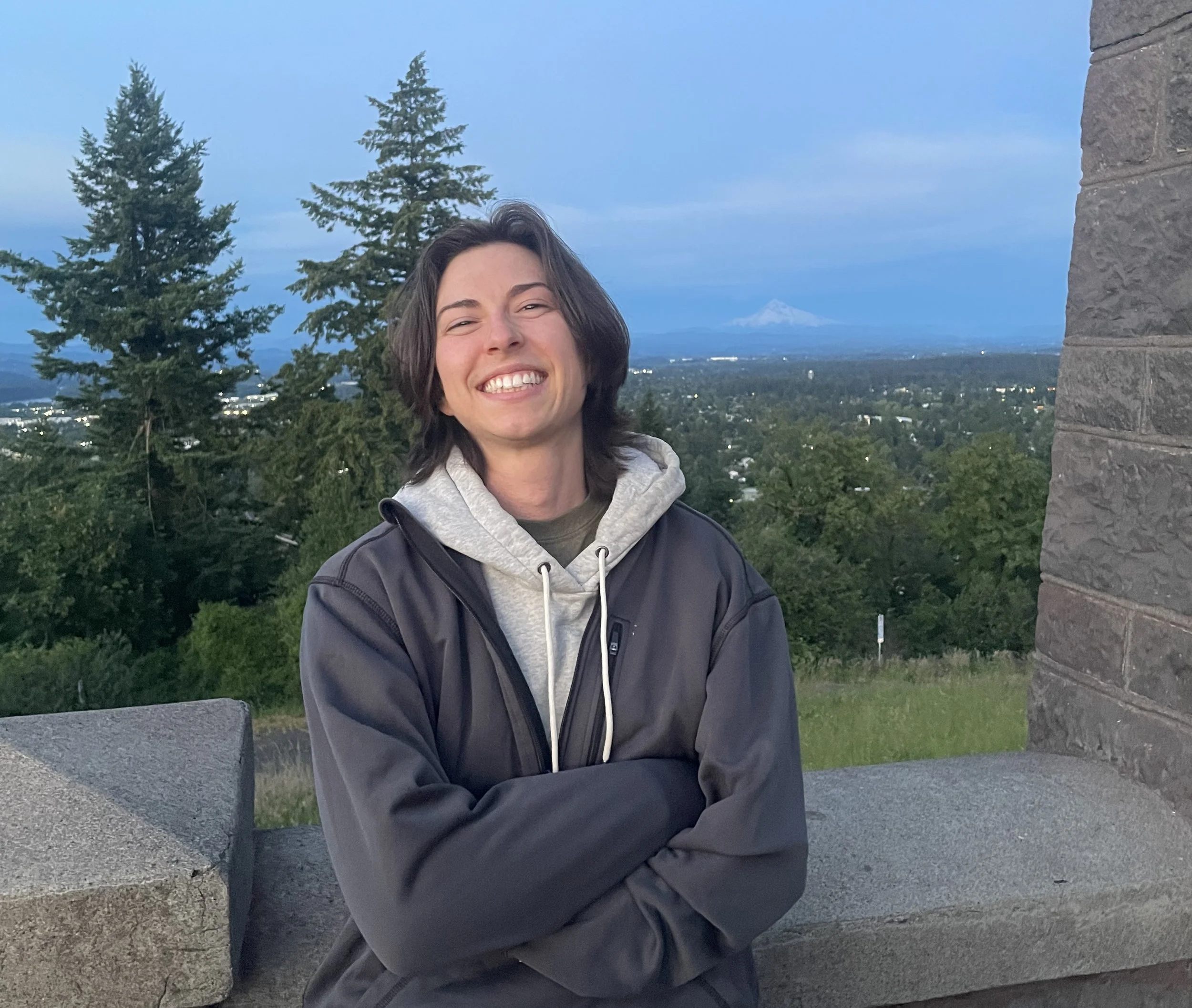 A smiling person with short dark hair standing outdoors near a stone wall, with trees and a distant mountain in the background.