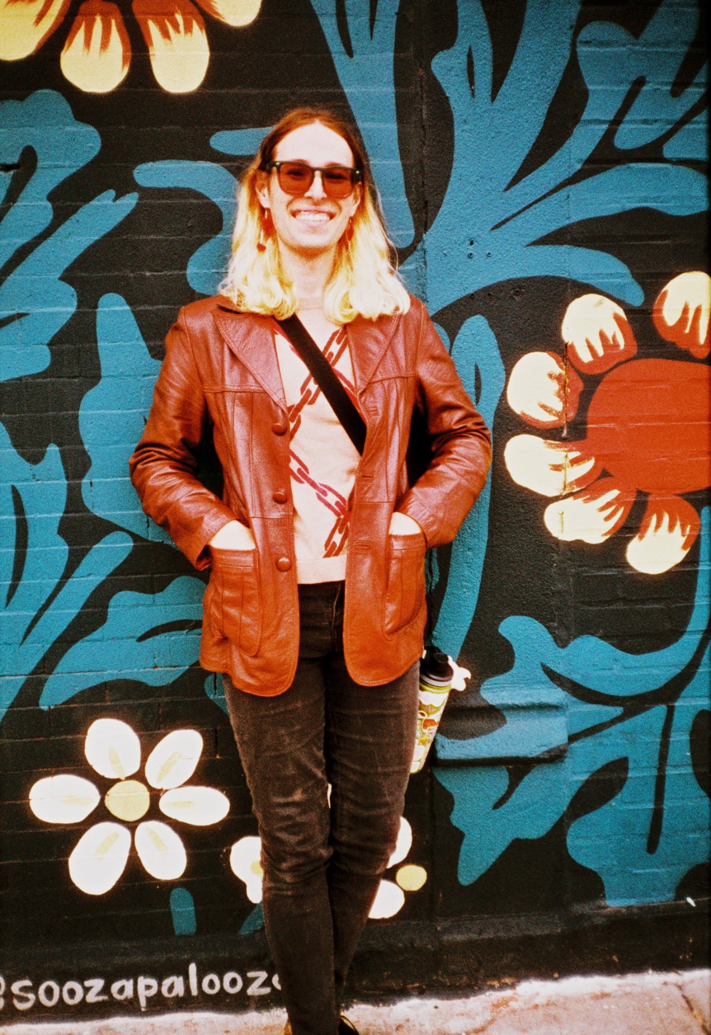 A person with blonde hair, wearing a brown leather jacket, sunglasses, and dark pants, standing in front of a colorful mural with floral and leaf patterns.