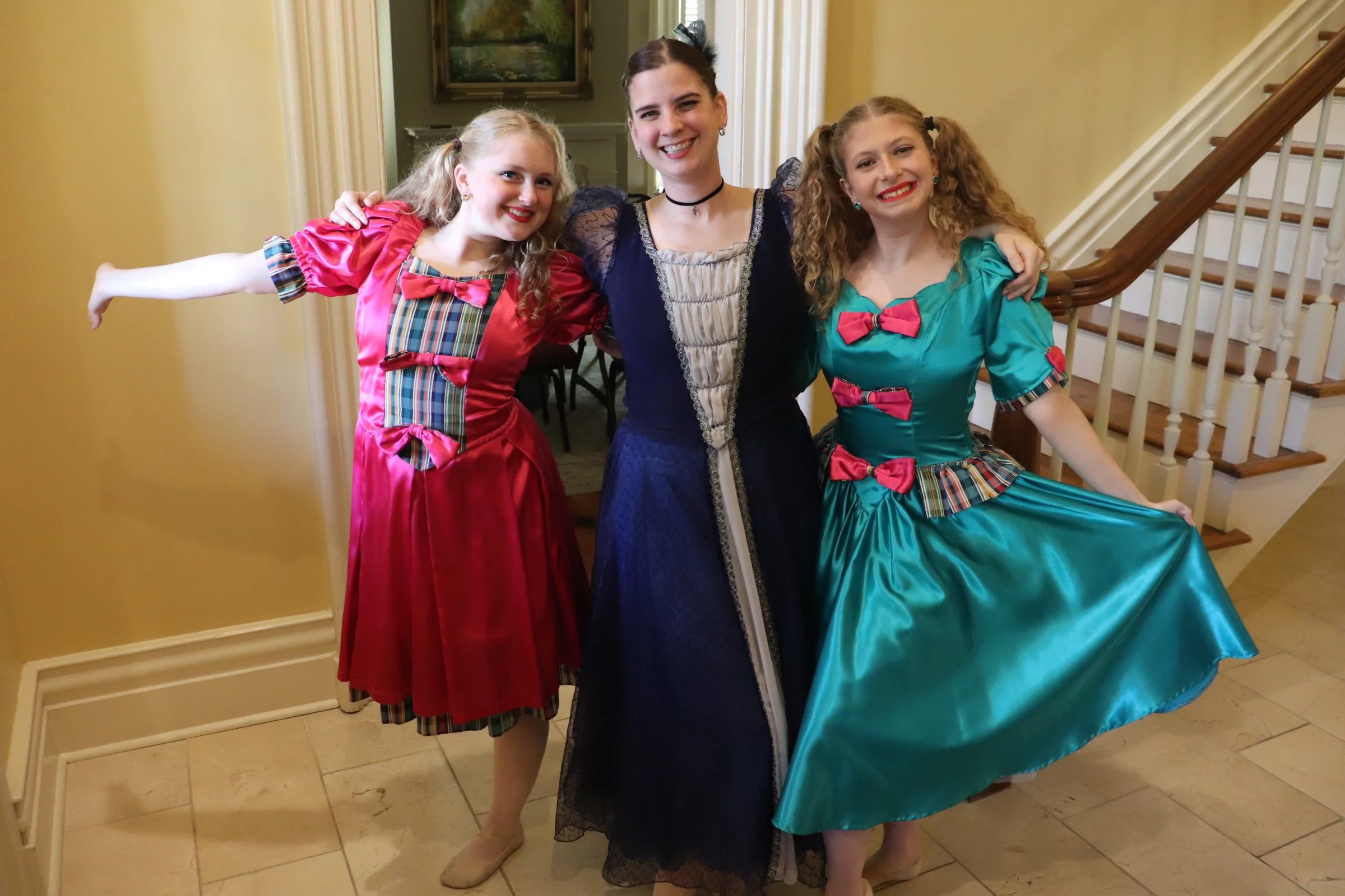 Three young women dressed in colorful, vintage-style dresses, standing together on a staircase inside a home. They are smiling and appear to be enjoying a special occasion or celebration.