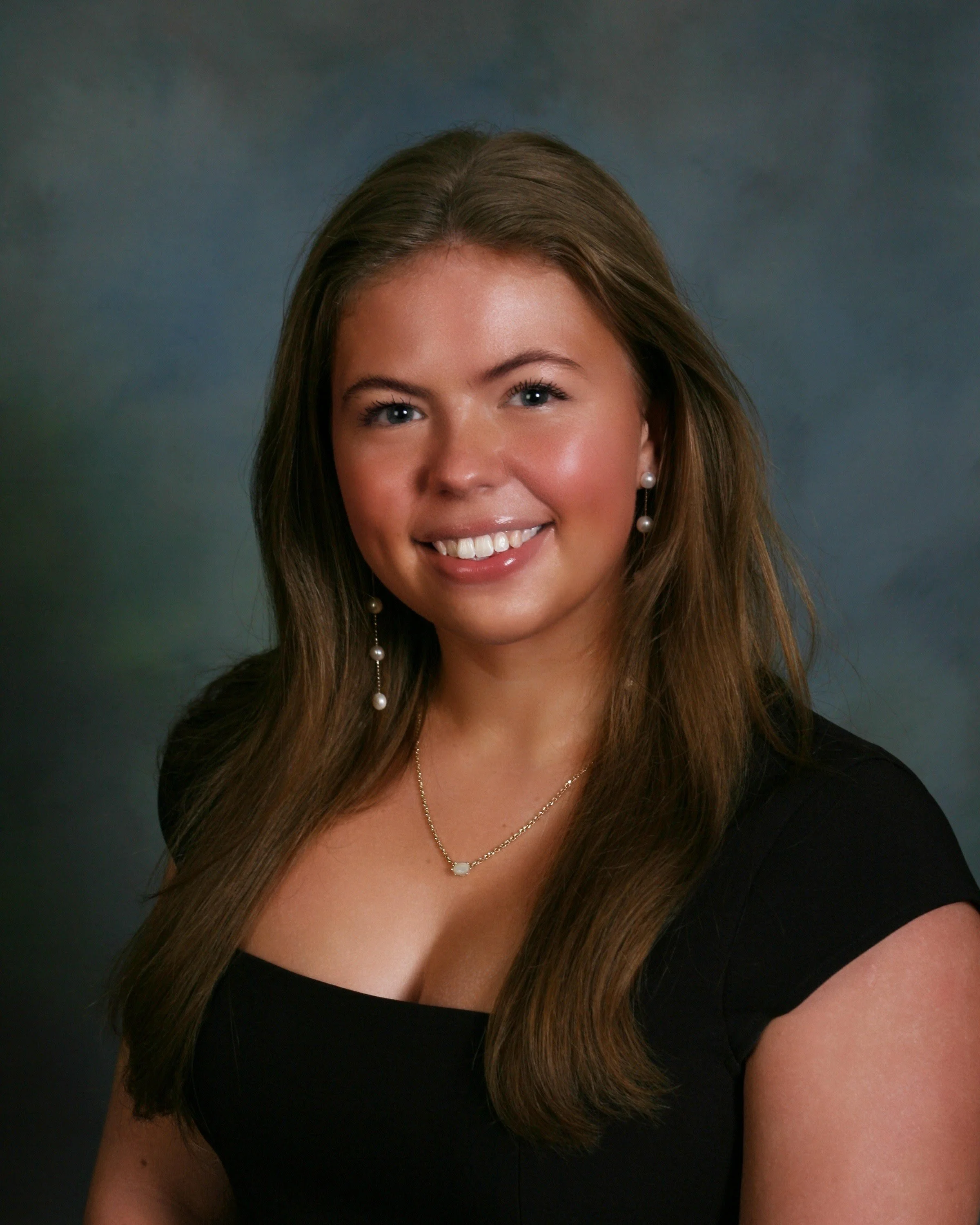 Close-up of a young woman with long brown hair, smiling, wearing pearl earrings and a delicate necklace, against a blurred dark background.