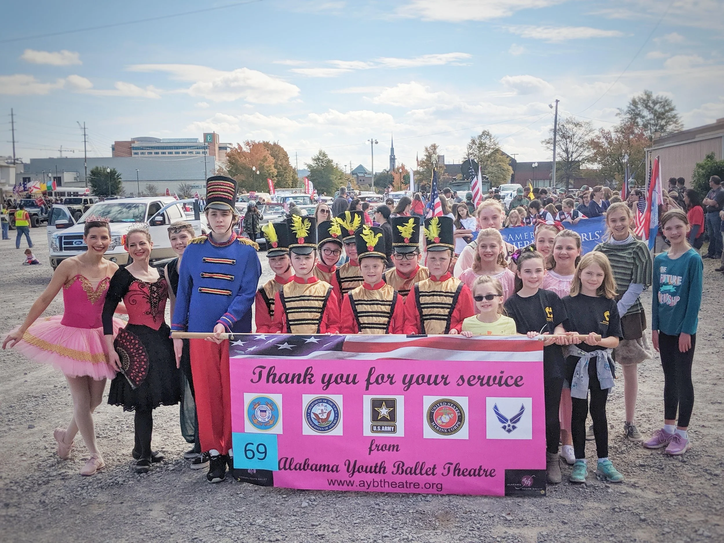 Group of young dancers and performers holding a pink banner that reads 'Thank you for your service' during a parade, with flags, marching bands, and spectators in the background on a bright day.