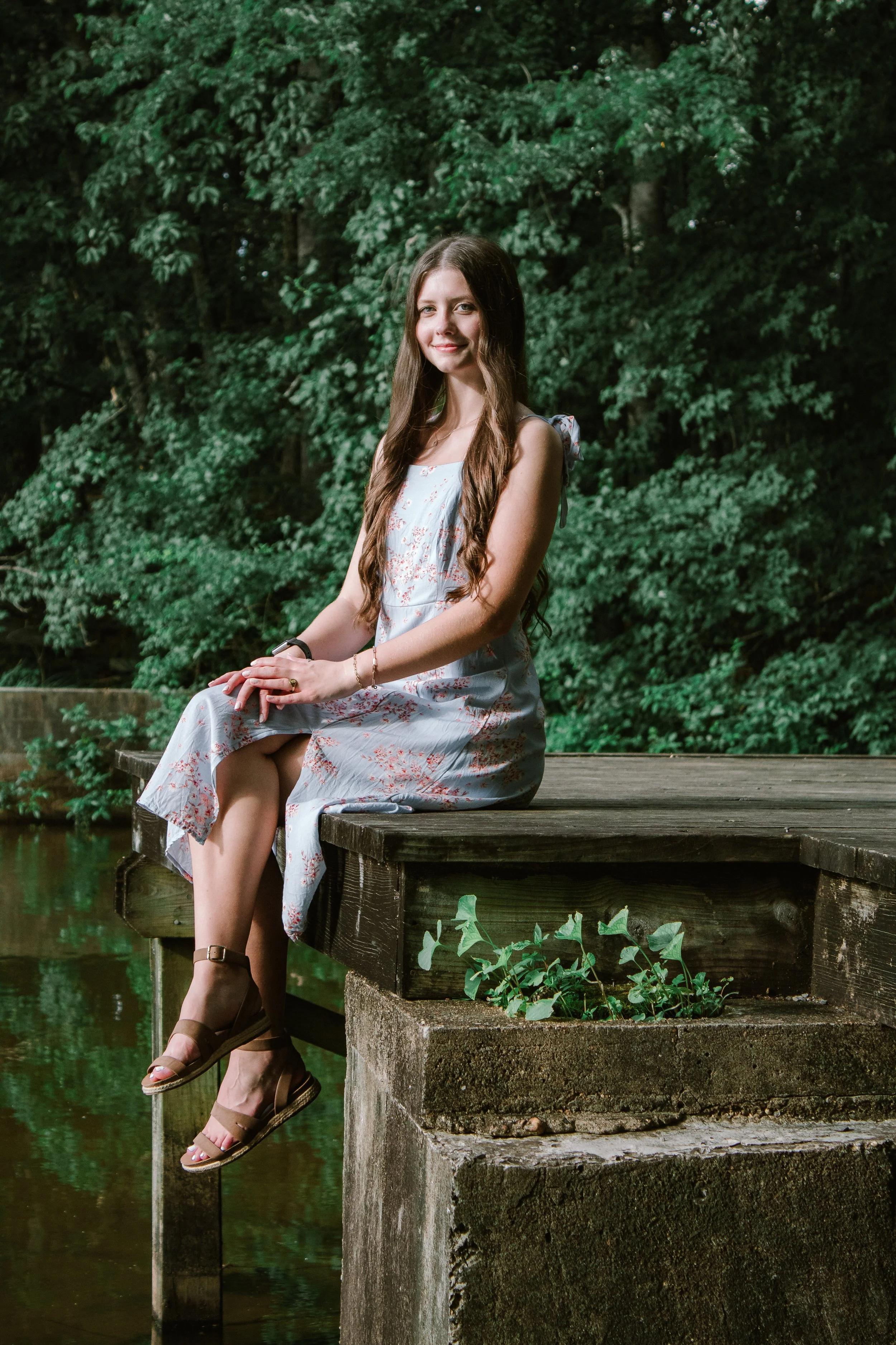 A young woman sitting on a wooden dock over water, surrounded by lush green trees, wearing a light-colored dress with floral patterns, brown sandals, and looking at the camera with a gentle smile.