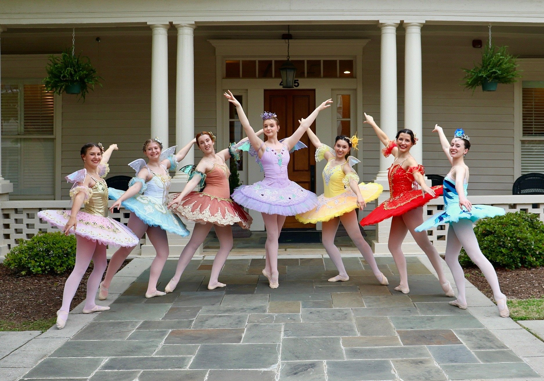 Eight ballet dancers in colorful tutus and tiaras performing a pose on a porch with columns, greenery, and a house in the background.