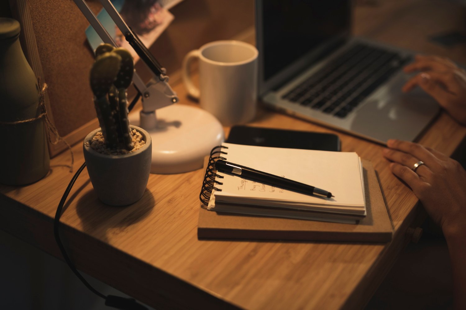 A cluttered wooden desk with a potted cactus, a white desk lamp, an open notebook with a pen, a white coffee mug, a laptop, and a smartphone, with hands typing on the laptop keyboard.