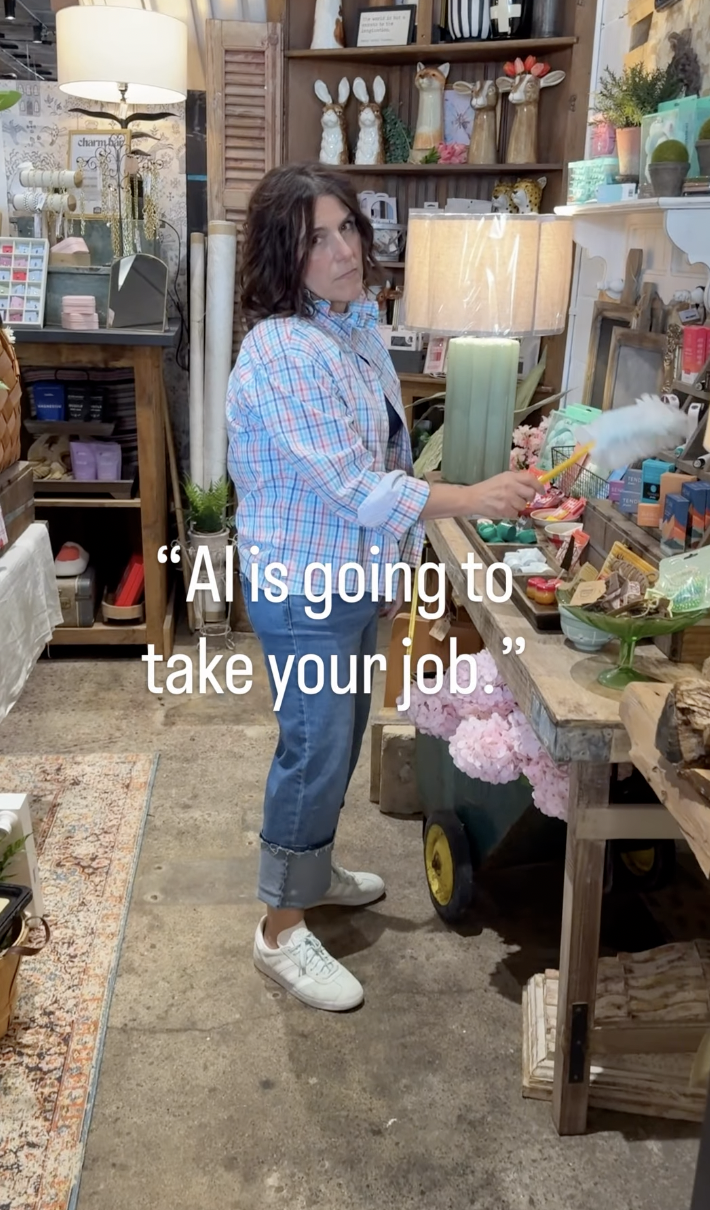 A woman with dark, wavy hair wearing a plaid shirt and jeans standing in a boutique store with home decor items. She is holding a duster and looking at the camera with a serious expression, surrounded by shelves of decorative objects, plants, and furniture. The quote on the image says, "AI is going to take your job."