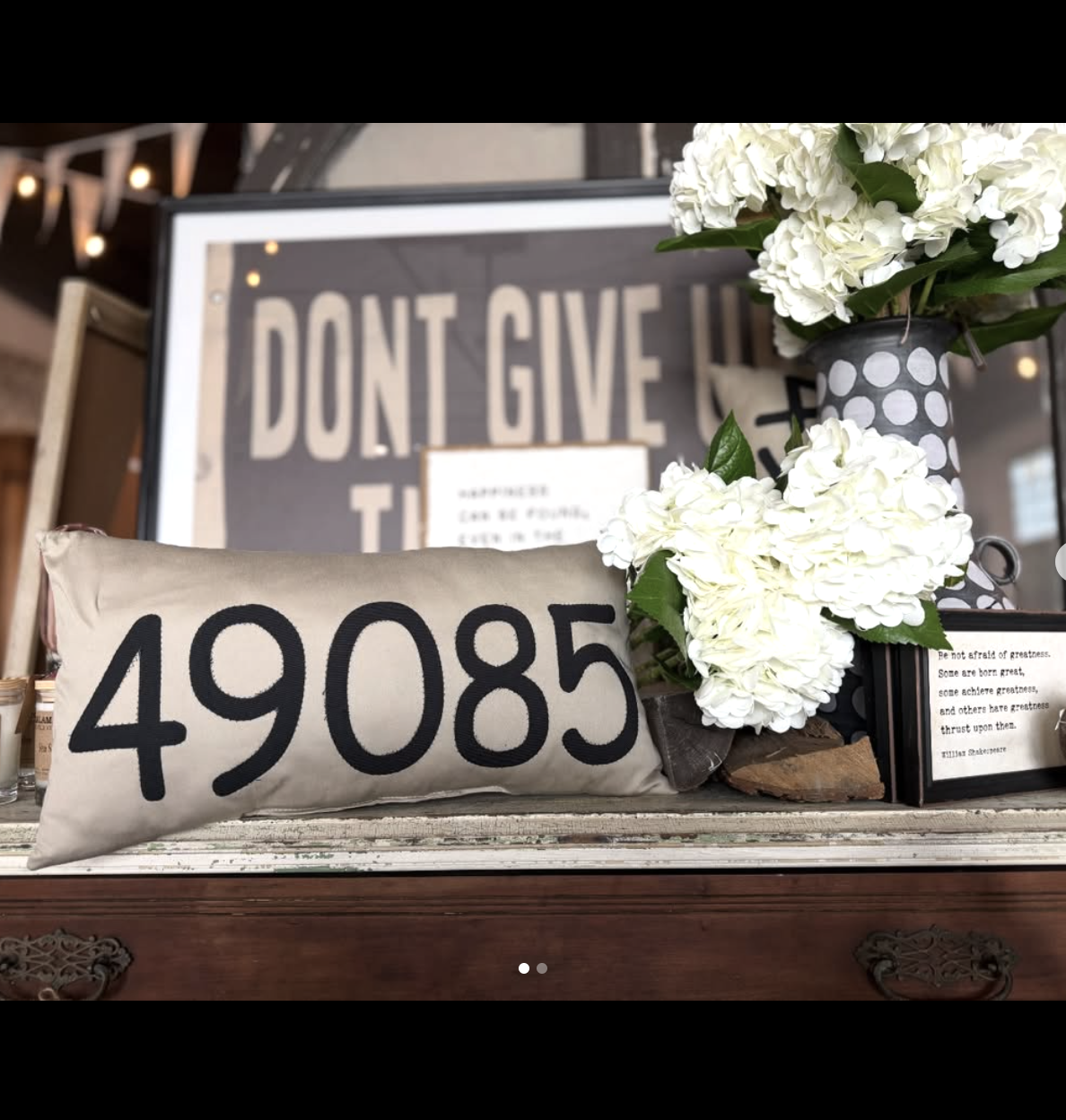 Decorative display with a beige pillow bearing the number 49085 next to a bouquet of white hydrangeas in a polka dot black and white vase, and a framed quote, with a background poster that reads 'Don't Give Up'.