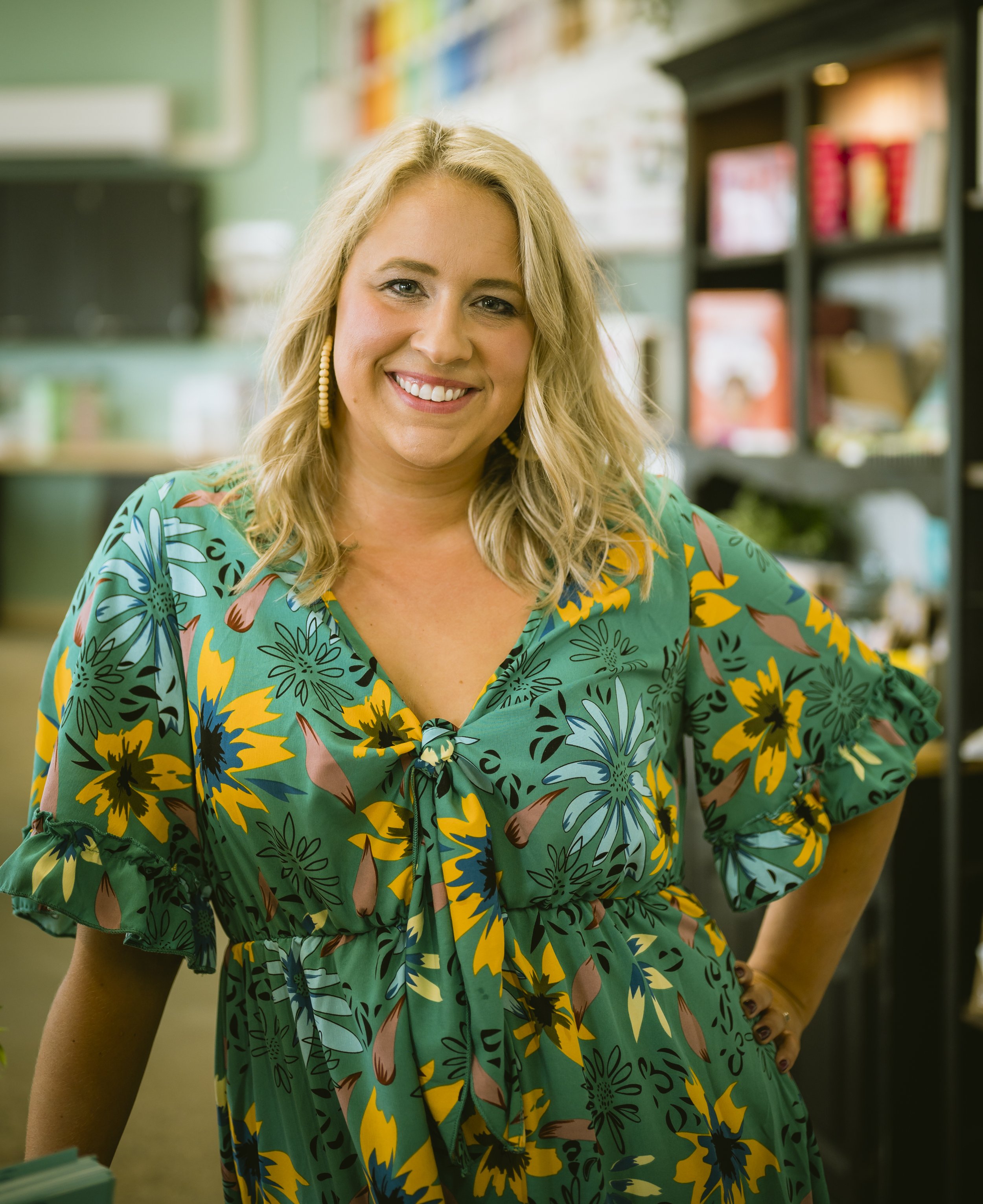 A smiling woman with blonde wavy hair wearing a colorful floral dress standing inside a room with bookshelves in the background.