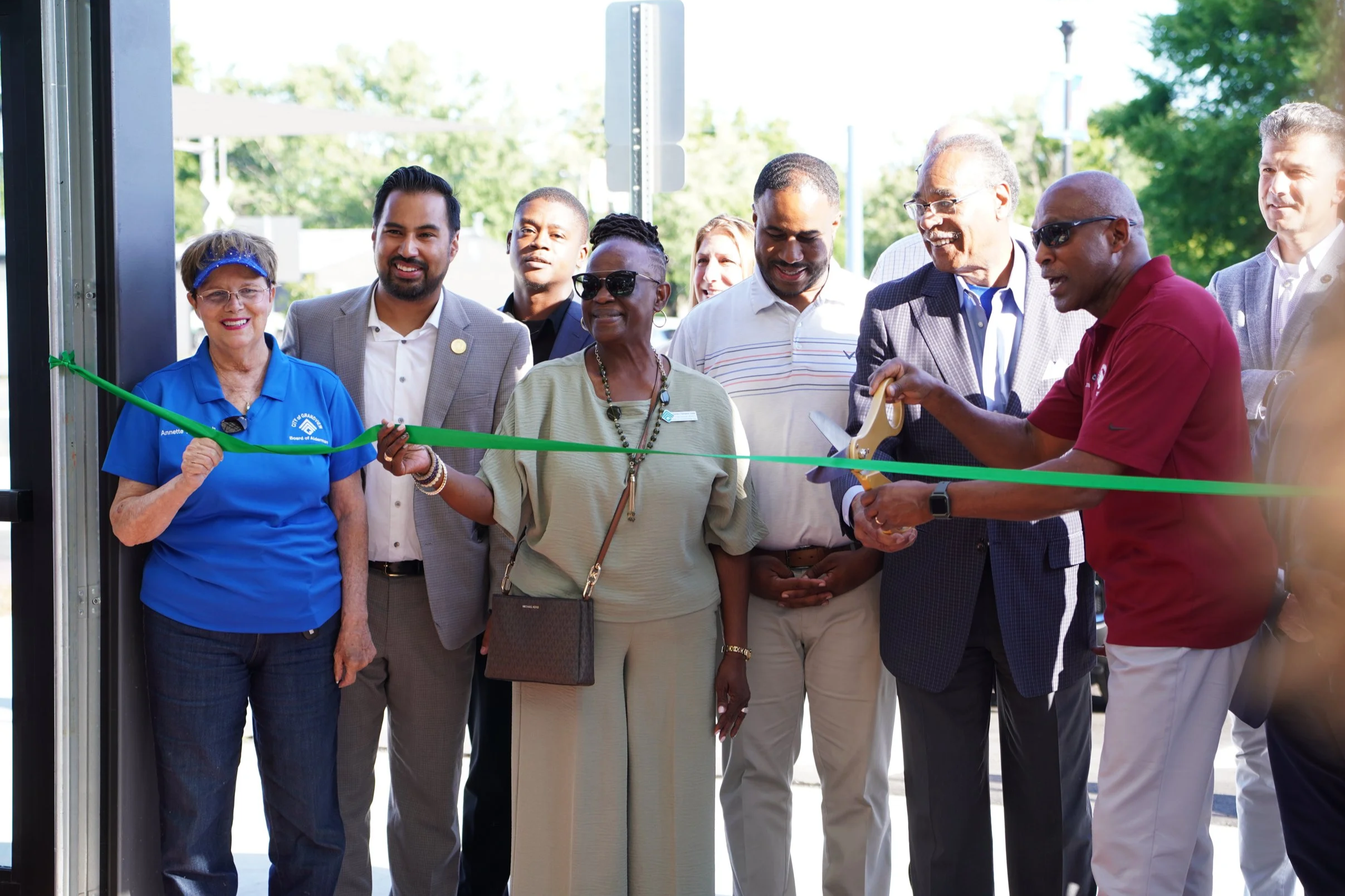 Group of diverse adults, including an older woman cutting a green ribbon, gathered outside for a ribbon-cutting ceremony, with some smiling and others observing.