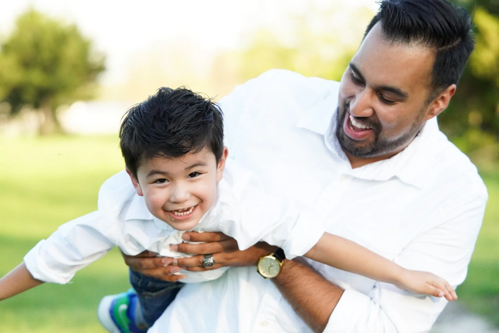 Man in white shirt holding young boy outdoors on green grass, both smiling and playing.