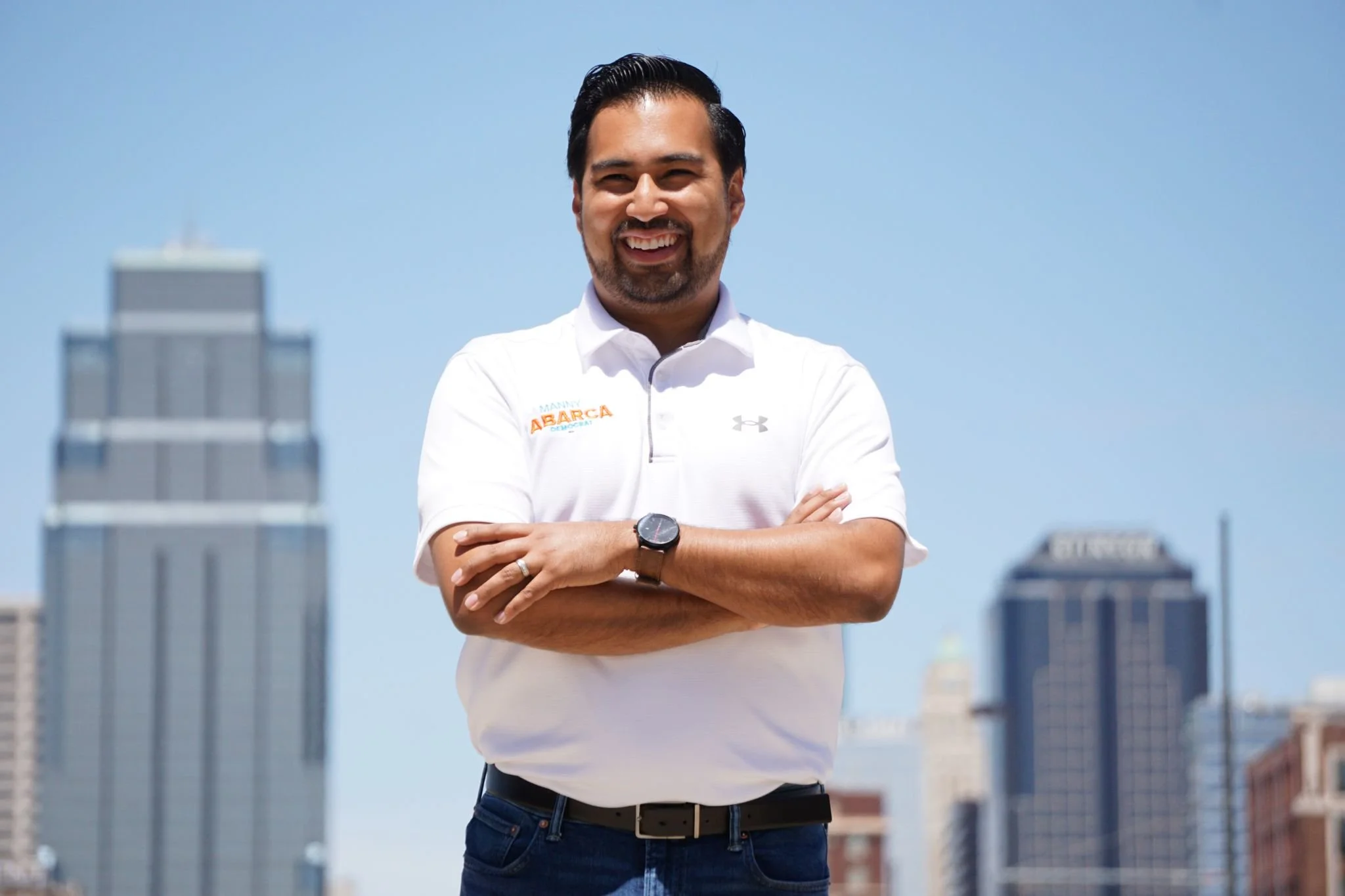 A man with dark hair and a beard smiling, wearing a white polo shirt with orange and blue text that says 'Manny ABARCA' and a watch, standing outdoors with a city skyline and blue sky in the background.