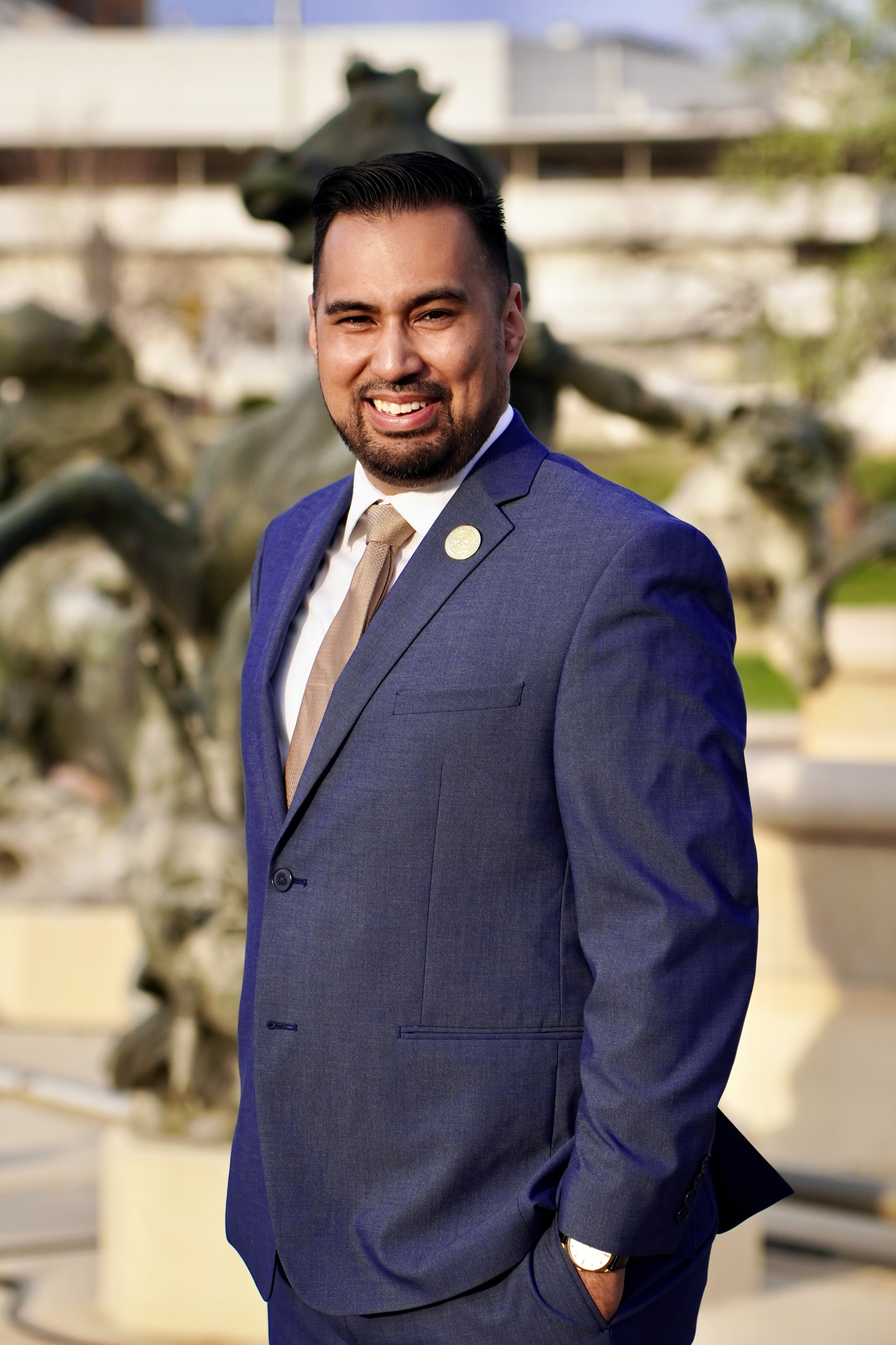A man in a blue suit with a light beige tie, white shirt, and gold watch standing outdoors in front of a sculpture of a horse.