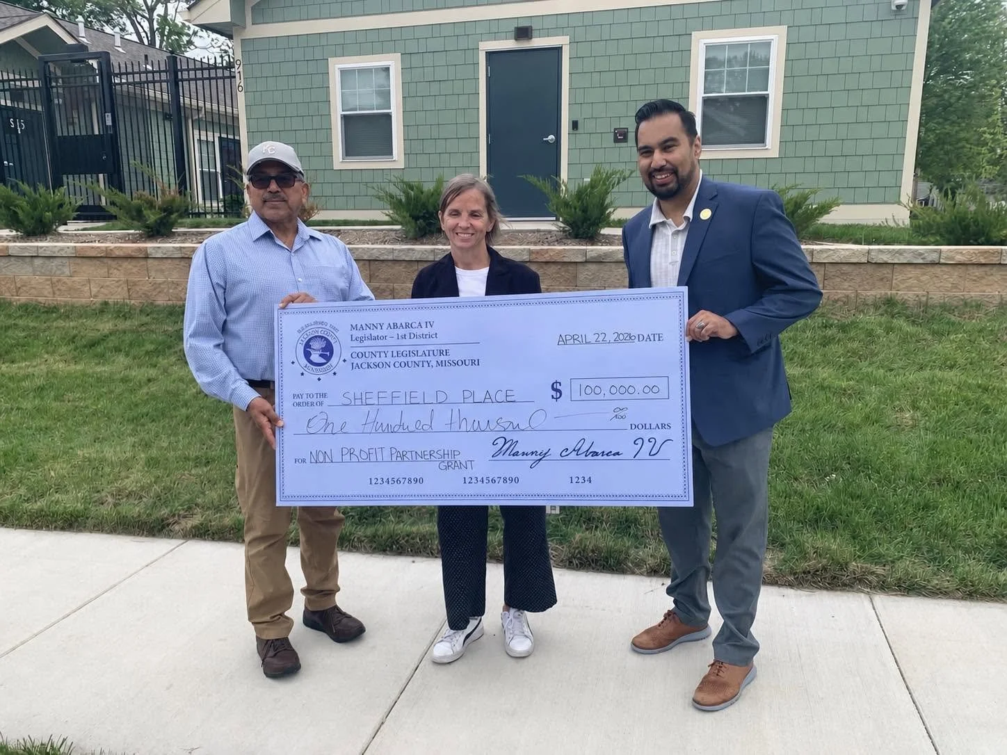 Three people standing outdoors in front of a house, holding an oversized check for $100,000 made out to Sheffield Place, for a nonprofit grant, dated April 22, 2026, smiling at the camera.