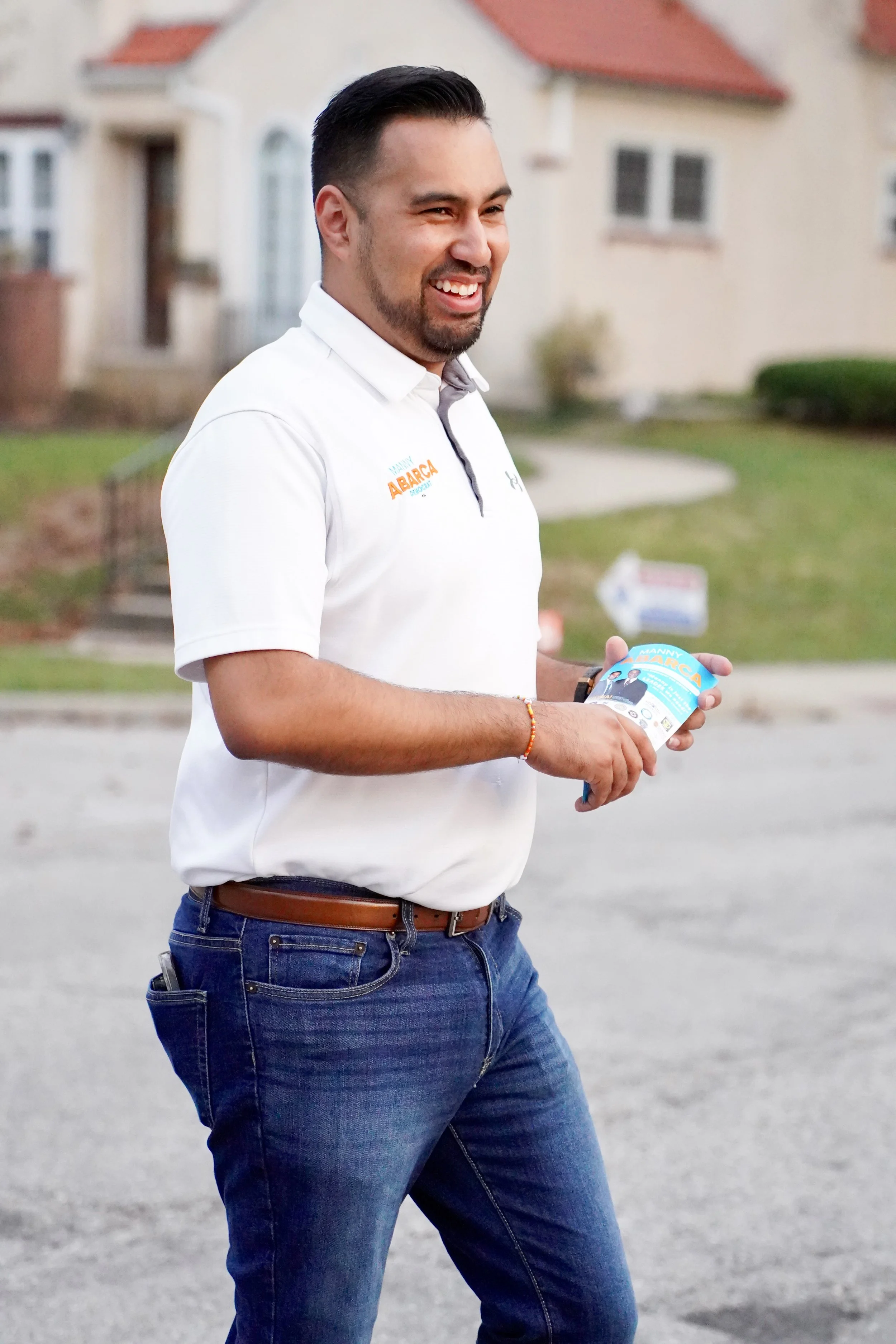 A man smiling outdoors, wearing a white polo shirt and jeans, holding flyers with campaign information, with residential houses in the background.