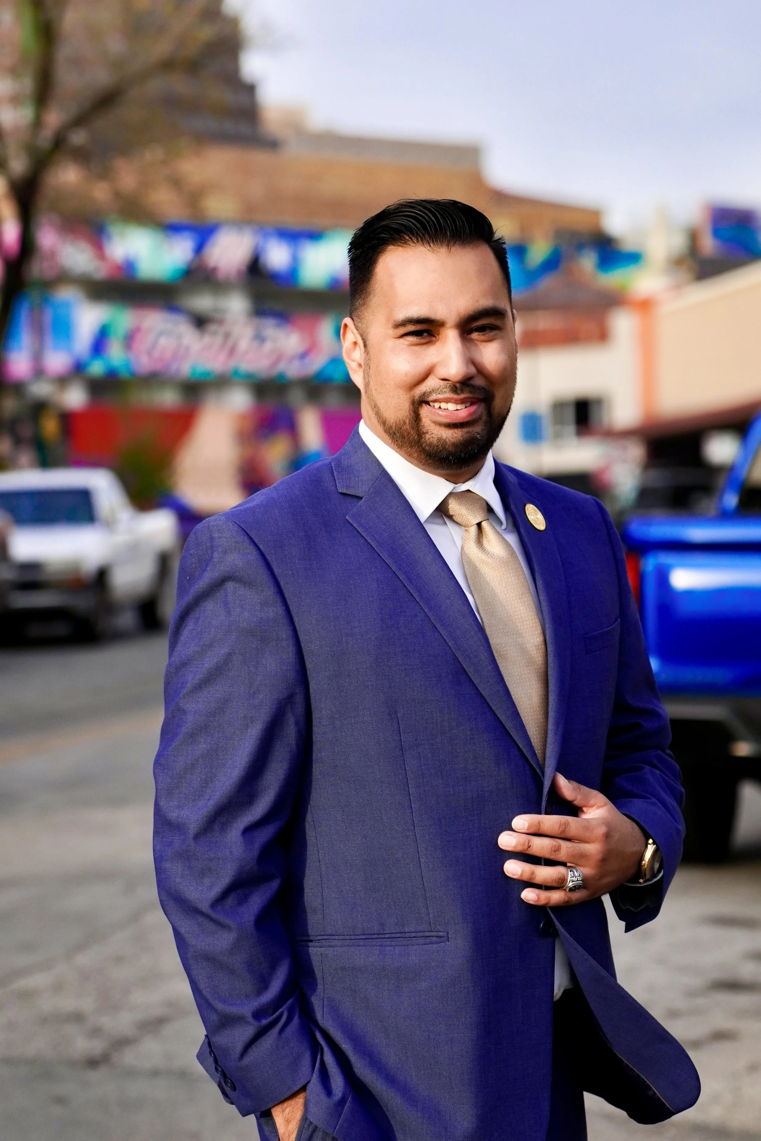 A man in a blue suit and tie standing outdoors with colorful murals and buildings in the background.