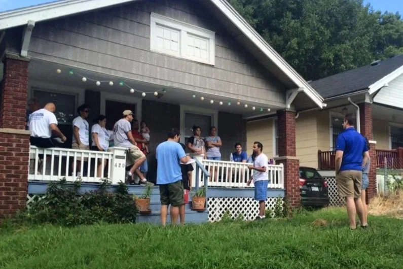 Group of people gather on a front porch of a house for a social event, some standing and some sitting, with a string of lights overhead and a small lawn in front.