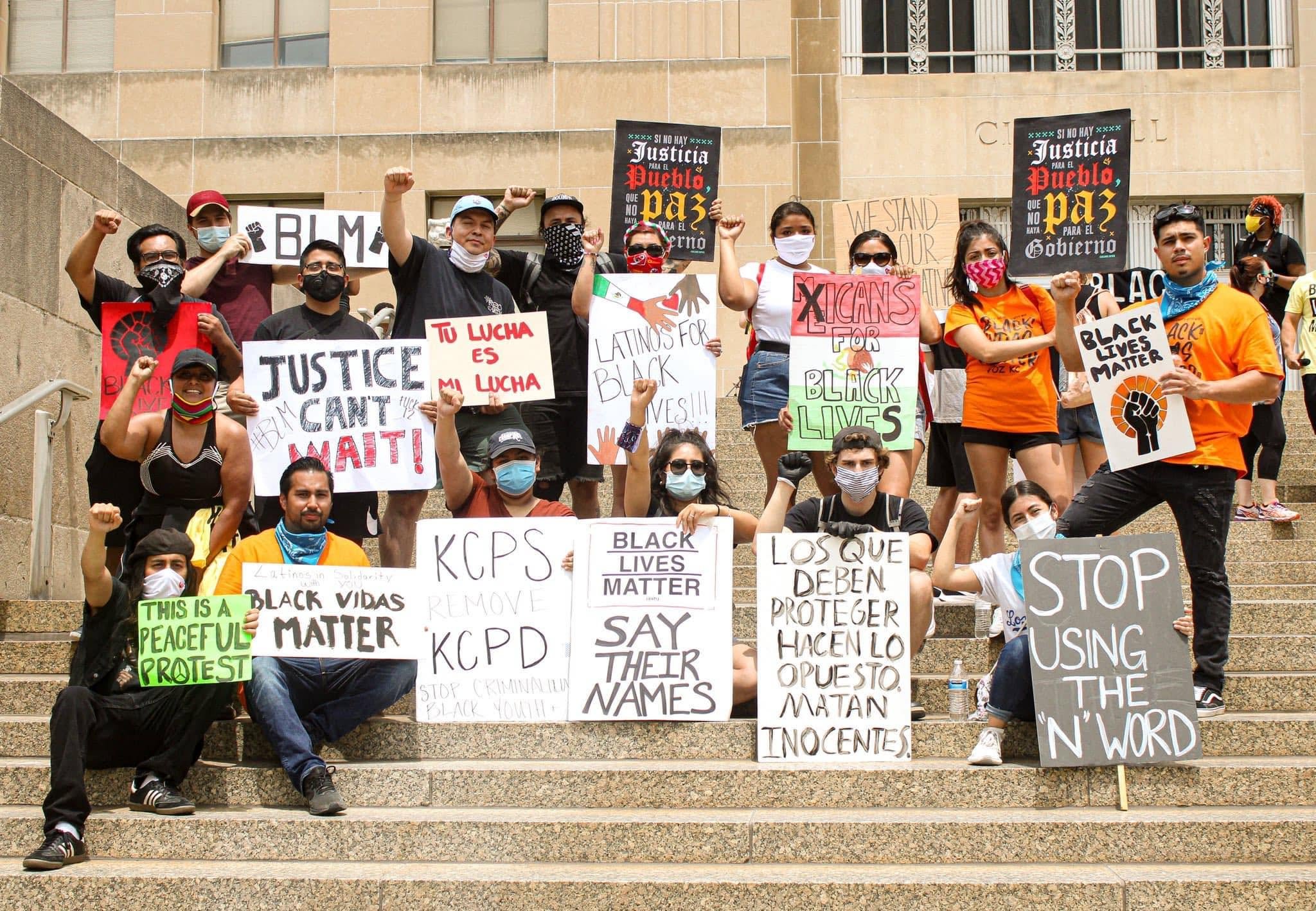 A group of people protesting on a set of outdoor stairs holding signs with messages about justice, Black Lives Matter, and anti-police sentiments. Some individuals are wearing masks and raising their fists.