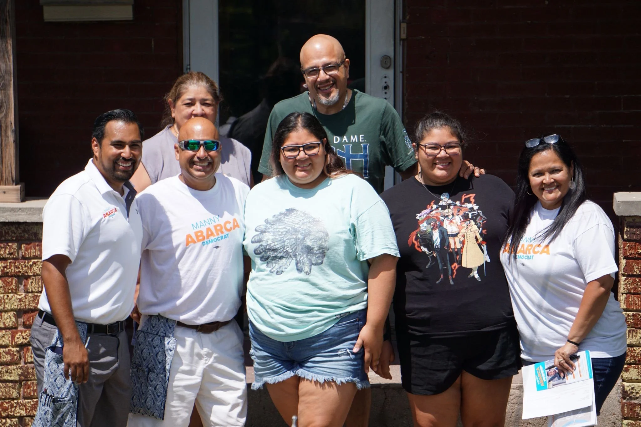 A group of seven smiling diverse adults standing outdoors in front of a brick and wood building, posing for a photo.