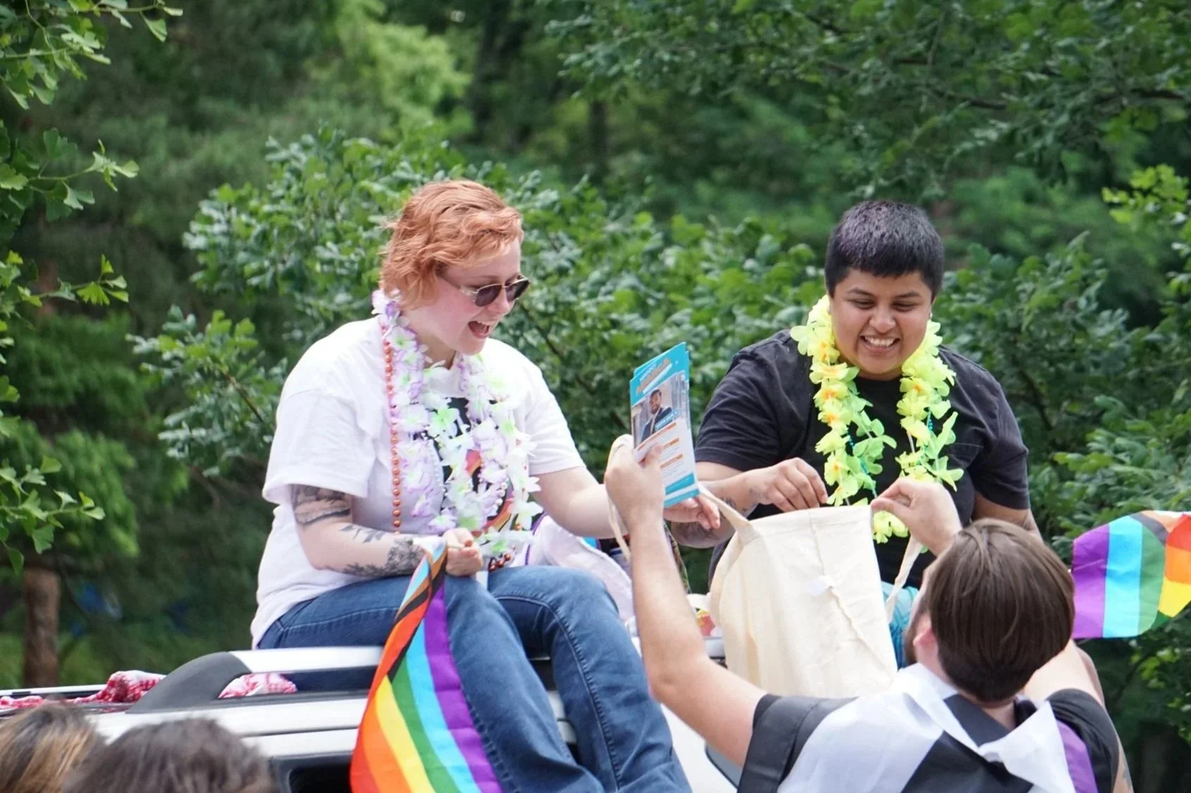 People celebrating at a parade or outdoor event, wearing leis and surrounded by rainbow flags, with trees in the background.