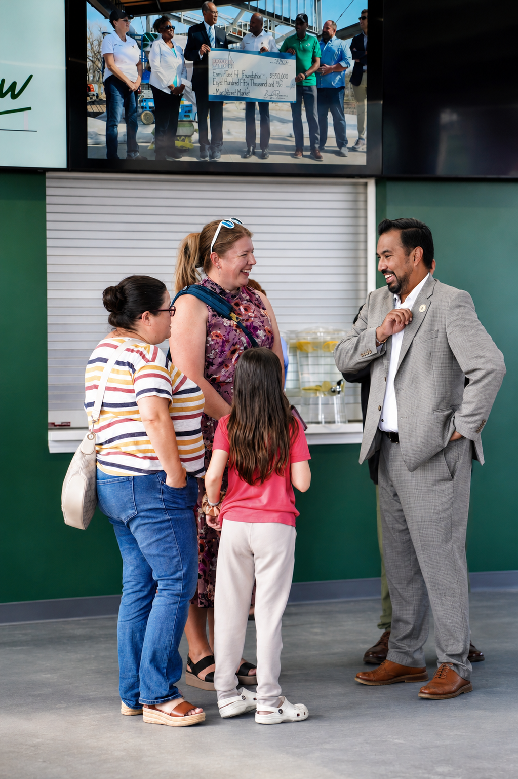 A group of four women, a man, and a young girl are talking and laughing inside a building with a green wall and a closed window. The women are wearing casual clothes; one has a striped shirt and another a floral dress. The man is in a light gray suit, and the girl is in a pink shirt and white pants. They seem happy and engaged in conversation.