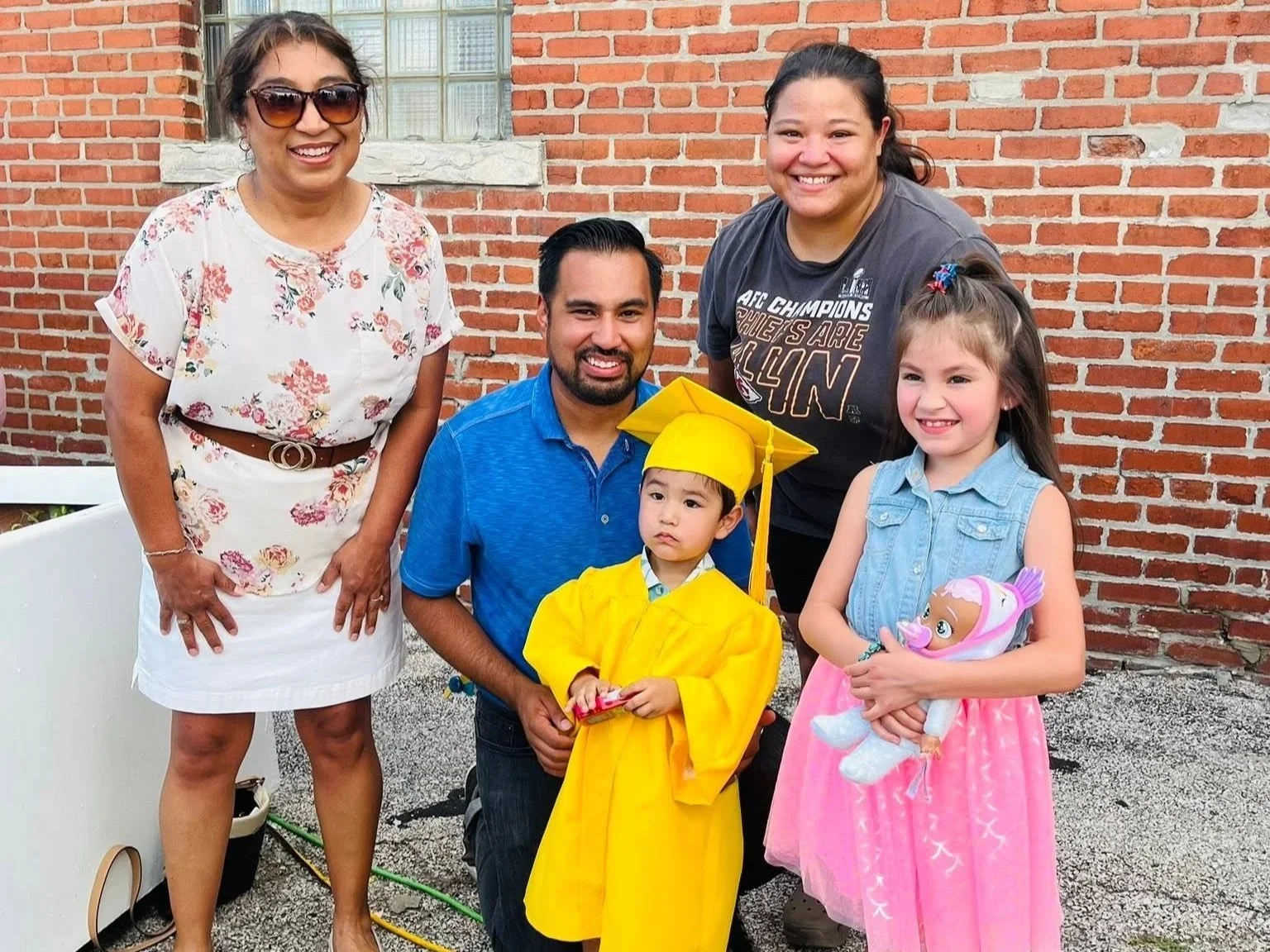 Group of five people, including a young boy in a yellow graduation gown and cap, standing in front of a brick wall outdoors, with two women and two girls smiling.