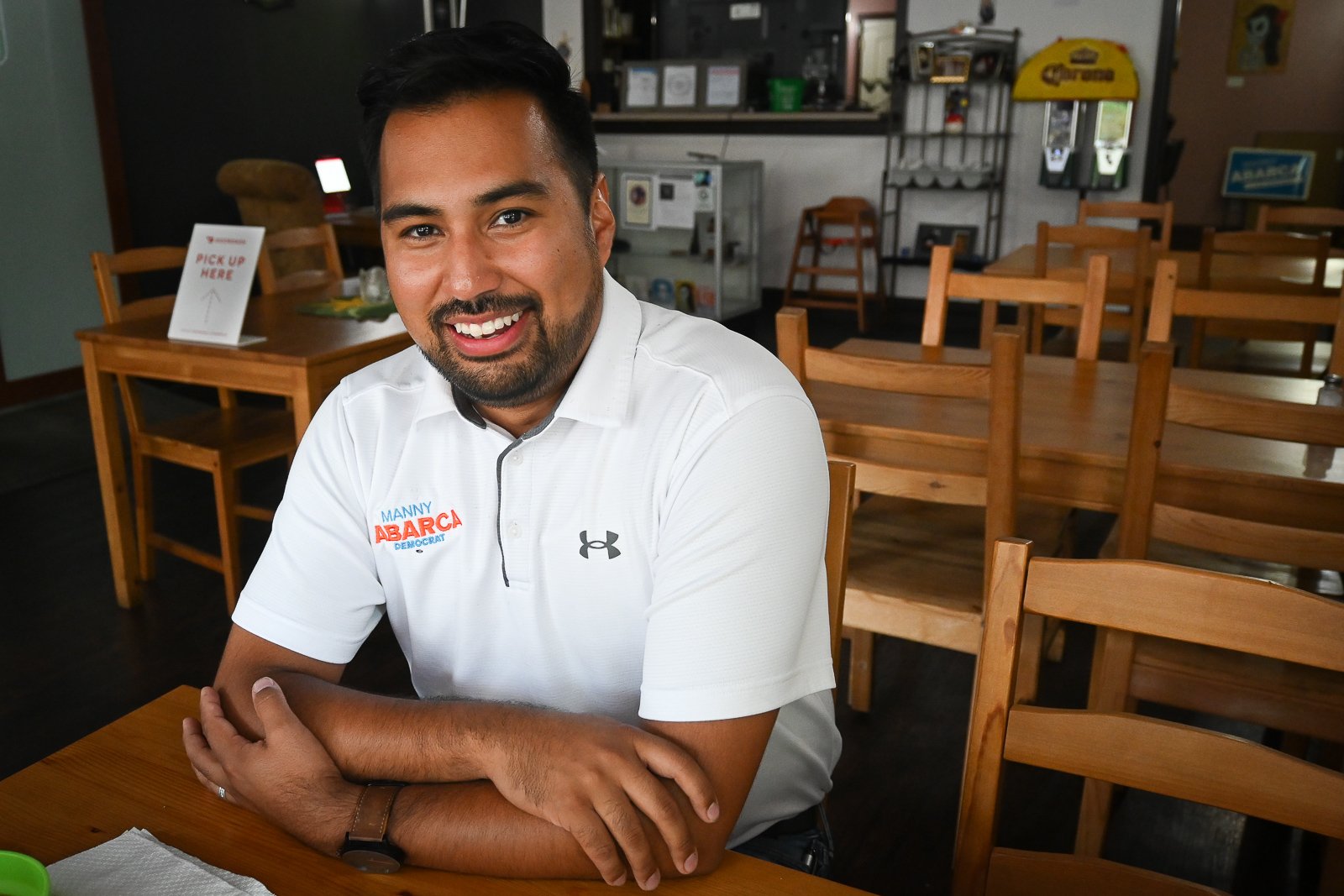 A smiling man with dark hair and a beard sitting at a wooden table in a restaurant, wearing a white polo shirt with political campaign logos.
