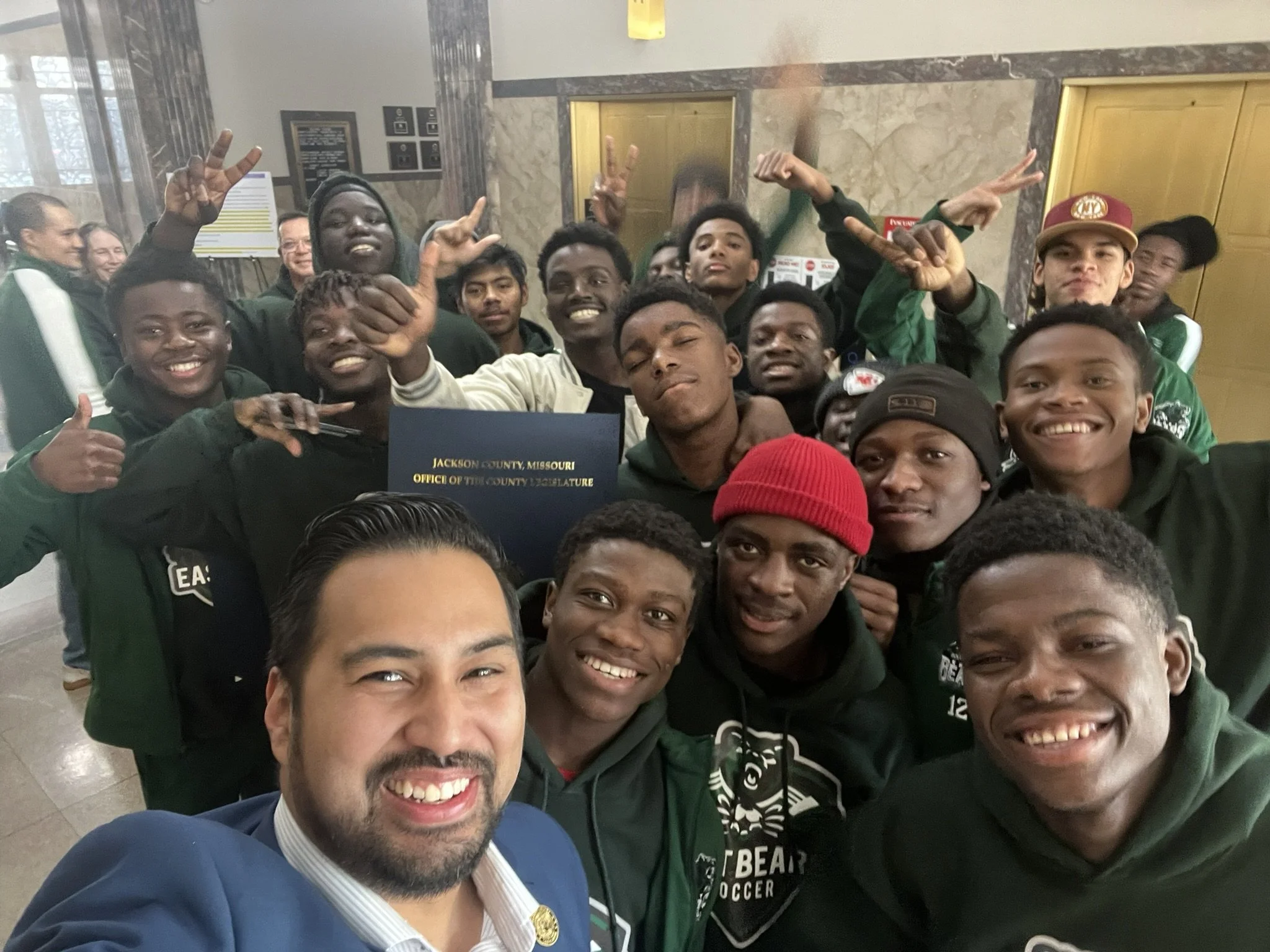 A group of young men and women taking a selfie together indoors, with a man in a suit in the foreground. The group is smiling and making peace signs. The background shows an elevator, some signs, and a marble wall. The badge on the man’s suit indicates they are at the Jackson County Office of the County Legislature in Missouri.