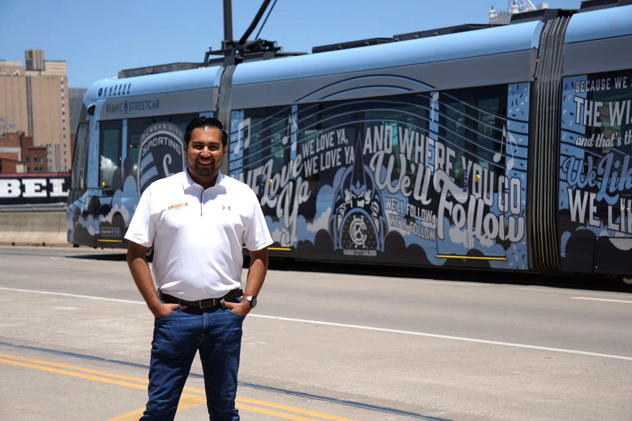 A man wearing a white polo shirt and jeans standing outdoors on a street with a parked electric bus behind him. The bus has a blue and black graphics design with text and music notes, and a cityscape in the background.