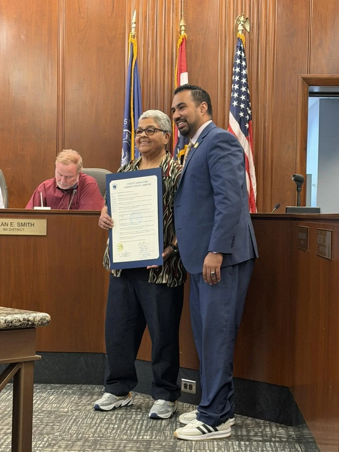 A woman and a man are standing together in a formal setting, holding a certificate. The woman is wearing glasses and a striped blouse, and the man is dressed in a blue suit. They are smiling. Behind them are American and state flags, and to the left, there is a seated man in a red shirt at a desk.