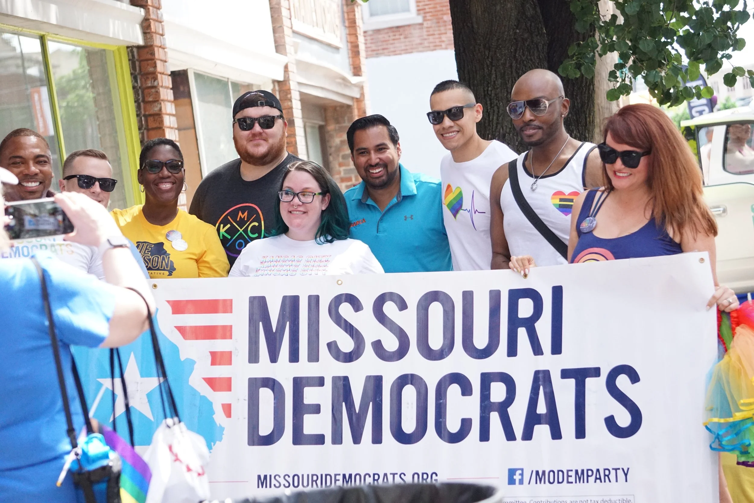 A diverse group of people at a pride parade holding a Missouri Democrats banner, some wearing rainbow-themed clothing and accessories, standing under a tree on a city street.