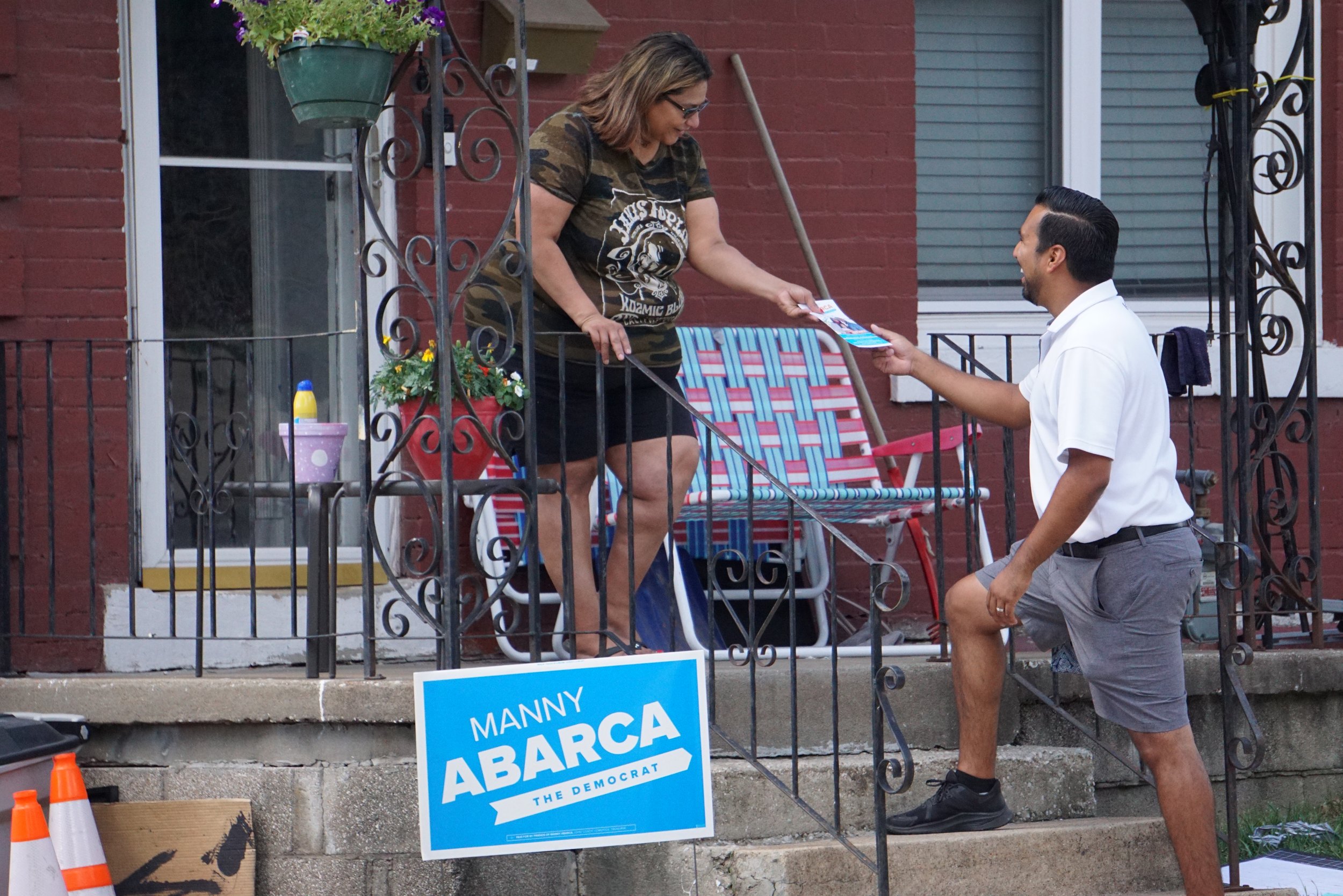 A woman on a porch handing a flyer to a man, with a campaign sign that reads 'Manny Abarca, The Democrat' visible in front of the porch.