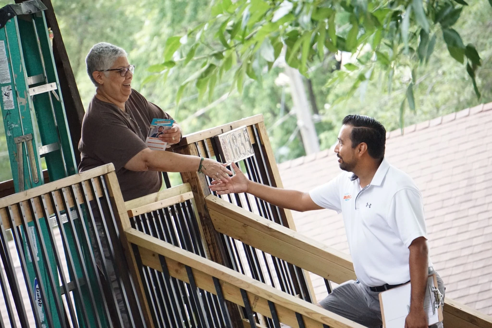 A smiling older woman with short gray hair and glasses, wearing a brown shirt, is shaking hands with a younger man with dark hair, a beard, and a white collared shirt. They are outdoors, next to a wooden bench and greenery.