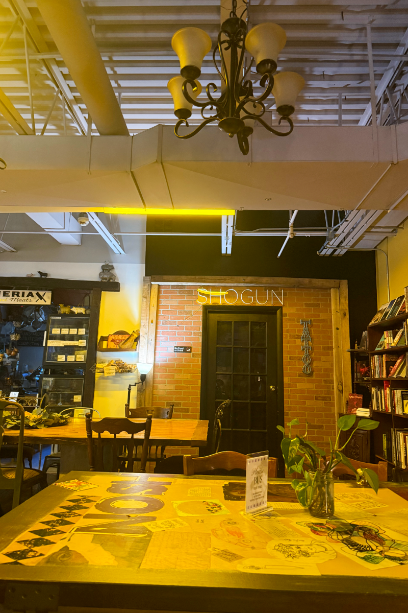 Wide view of Urban Food Brood with wooden tables, bookshelves, warm yellow lighting, a chandelier, and a brick wall with a SHOGUN sign.