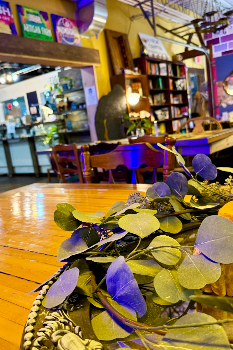 Close-up of greenery on a wooden table inside Urban Food Brood, with chairs, bookshelves, and colorful lighting blurred in the background.