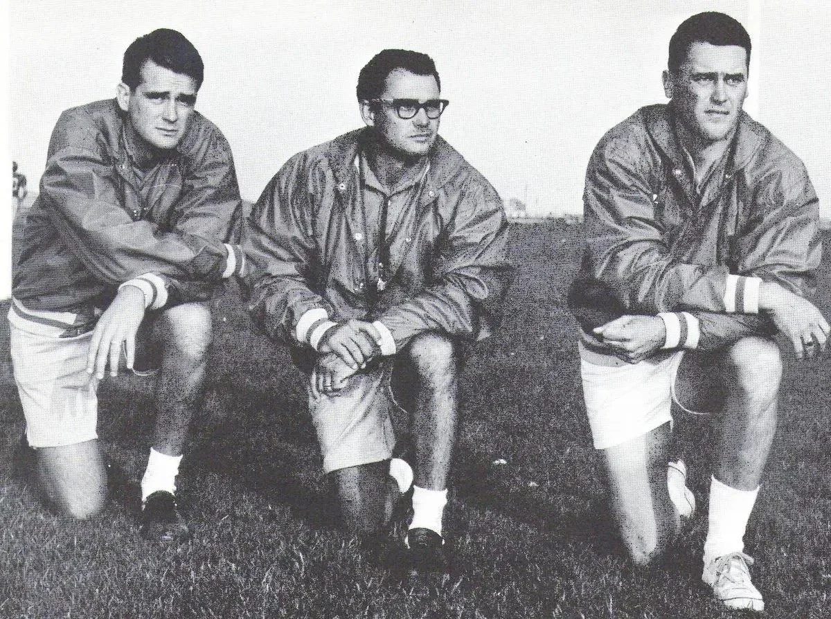 Black and white photo of three young men in vintage sports jackets, shorts, and knee-high socks crouching on a grassy field. Bruce Pickford.