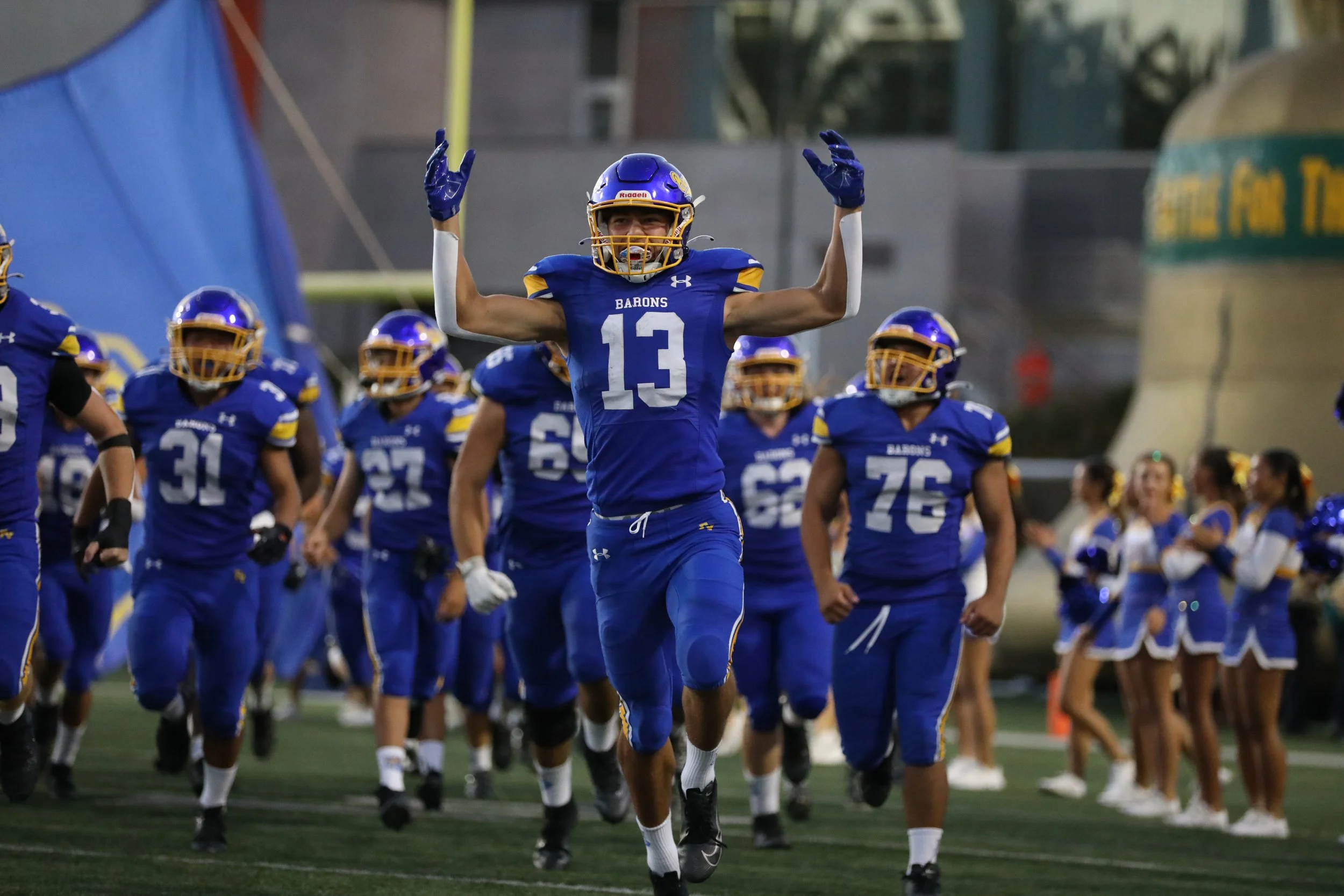 A group of football players in blue and yellow uniforms running onto the field under stadium lights, led by a player with the number 13 jersey raising his arms in celebration.