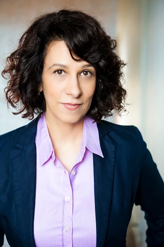 A woman with shoulder-length curly dark hair, wearing a purple collared shirt and a dark blazer, looking confidently at the camera.