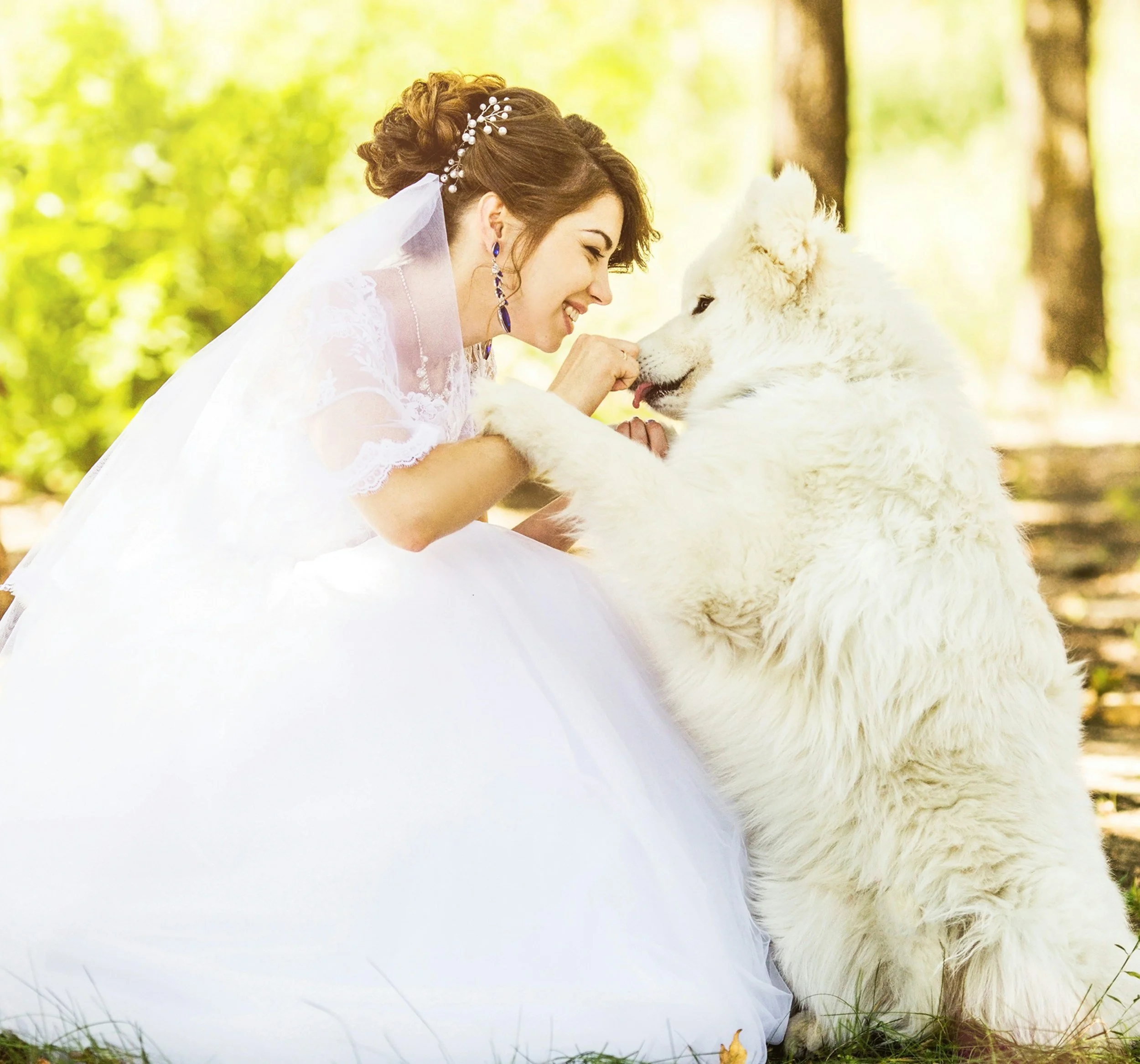 A woman in a white wedding dress with lace details, wearing jewelry including earrings and a hairpiece, smiling as she leans close to and touches the nose of a large white fluffy dog. They are outdoors in a wooded area with sunlight filtering through trees.