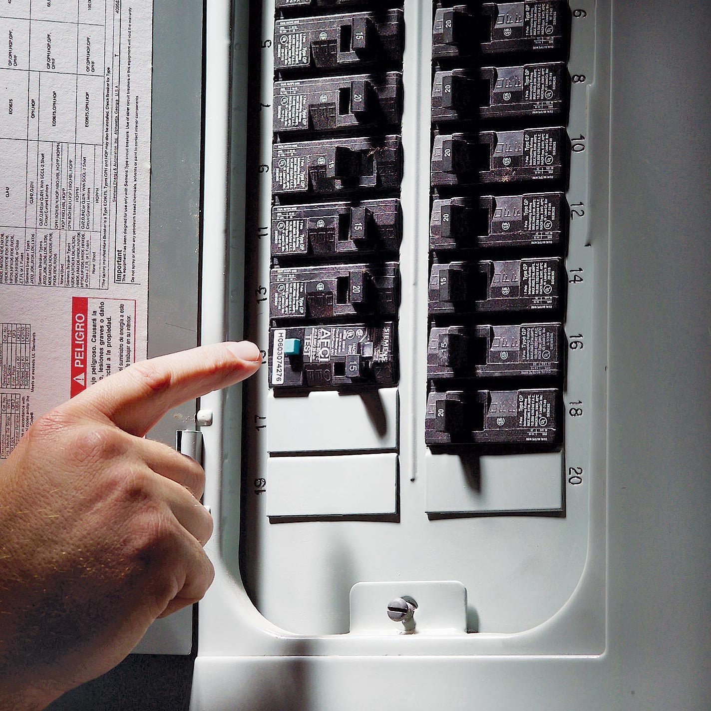 Close-up of a hand pointing to a circuit breaker in an electrical panel, with multiple black circuit breakers and a gray background.