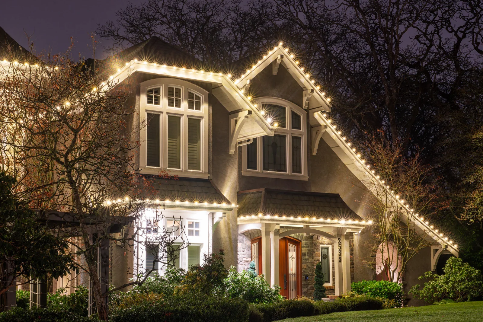 A large house decorated with string lights along the rooflines at dusk or night, surrounded by trees and shrubbery.