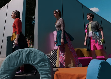 Trois jeunes femmes debout devant un bâtiment moderne, portant des vêtements colorés, avec des meubles d'extérieur en avant-plan.